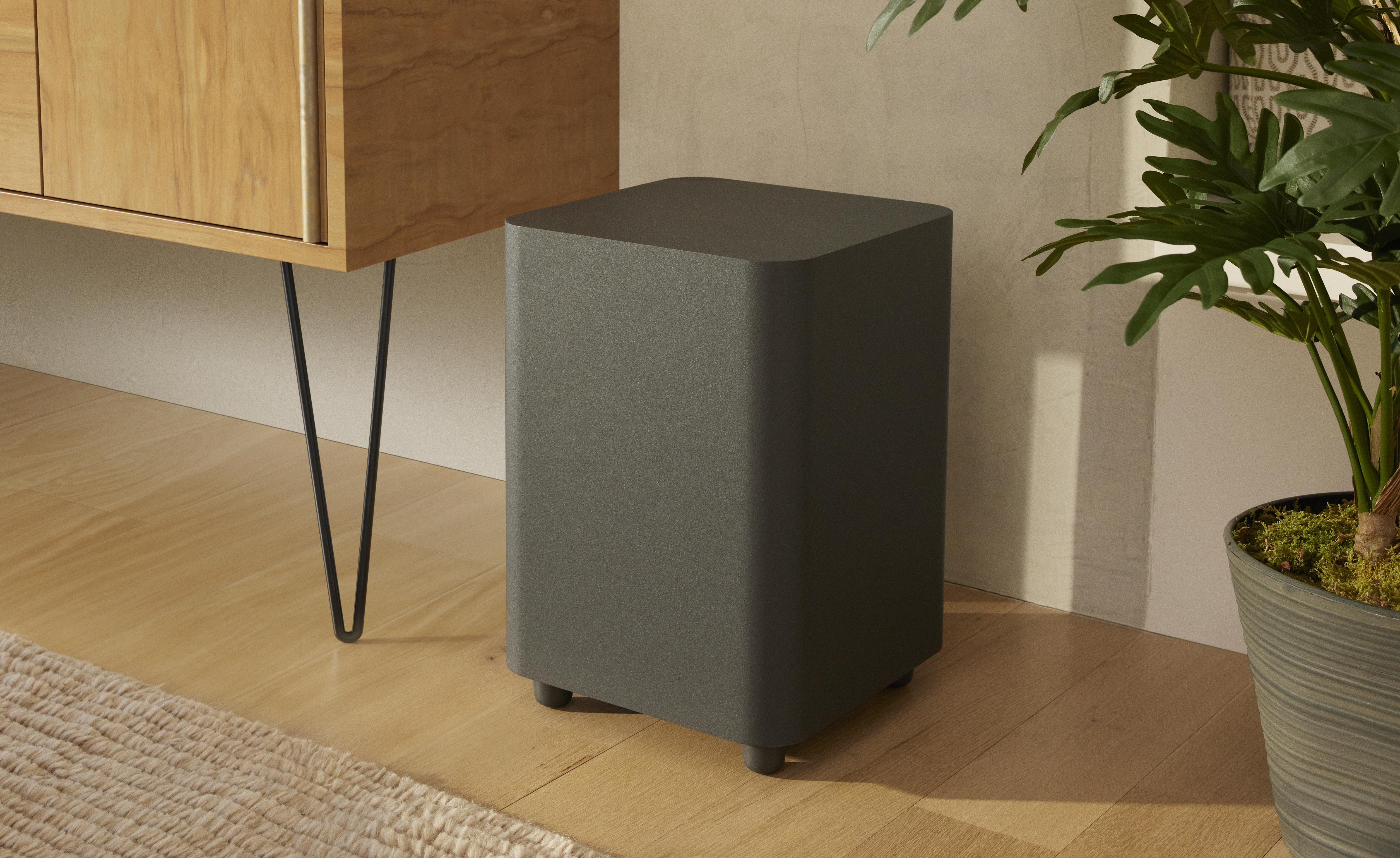 Ein grauer, quadratischer Subwoofer in einer Raumsituation. Auf Holzfußboden platziert, neben einer Pflanze.
