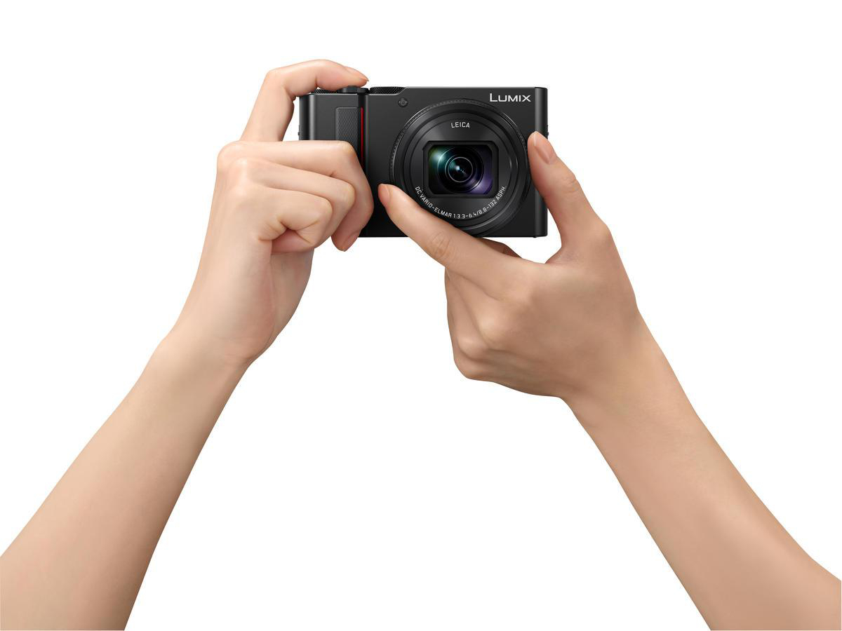 Eine Person hält eine schwarze Kamera und macht mit beiden Händen ein Foto. Weißer Hintergrund.
