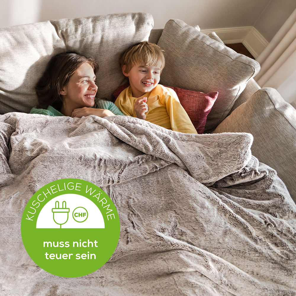Donna e bambino su un divano, coperti da una coperta. Testo: 'Kuschelige Wärme'.