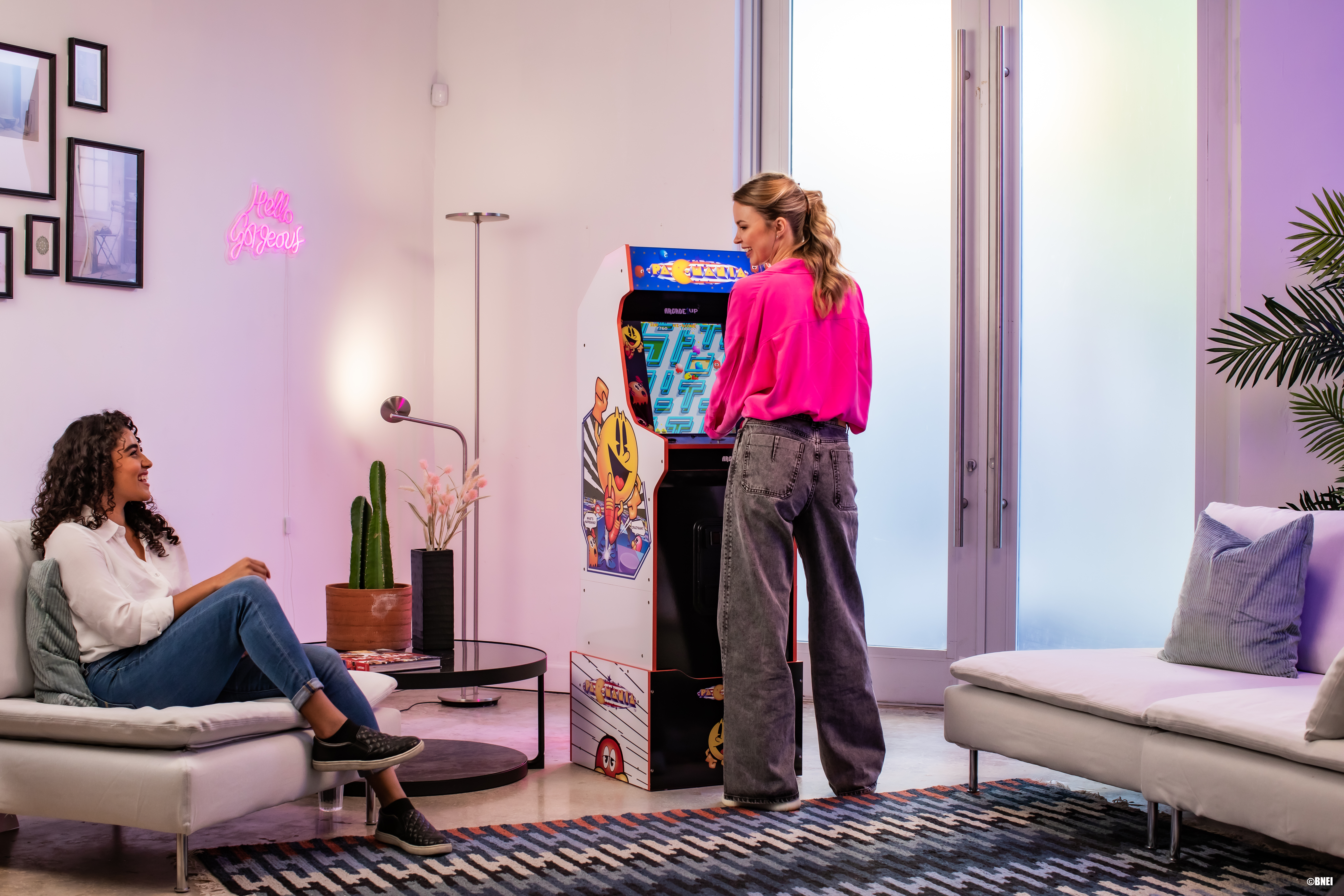 Zwei Frauen spielen ein Arcade-Spiel. Neon-Schild sagt "hello sweetie". Der Raum ist bunt.