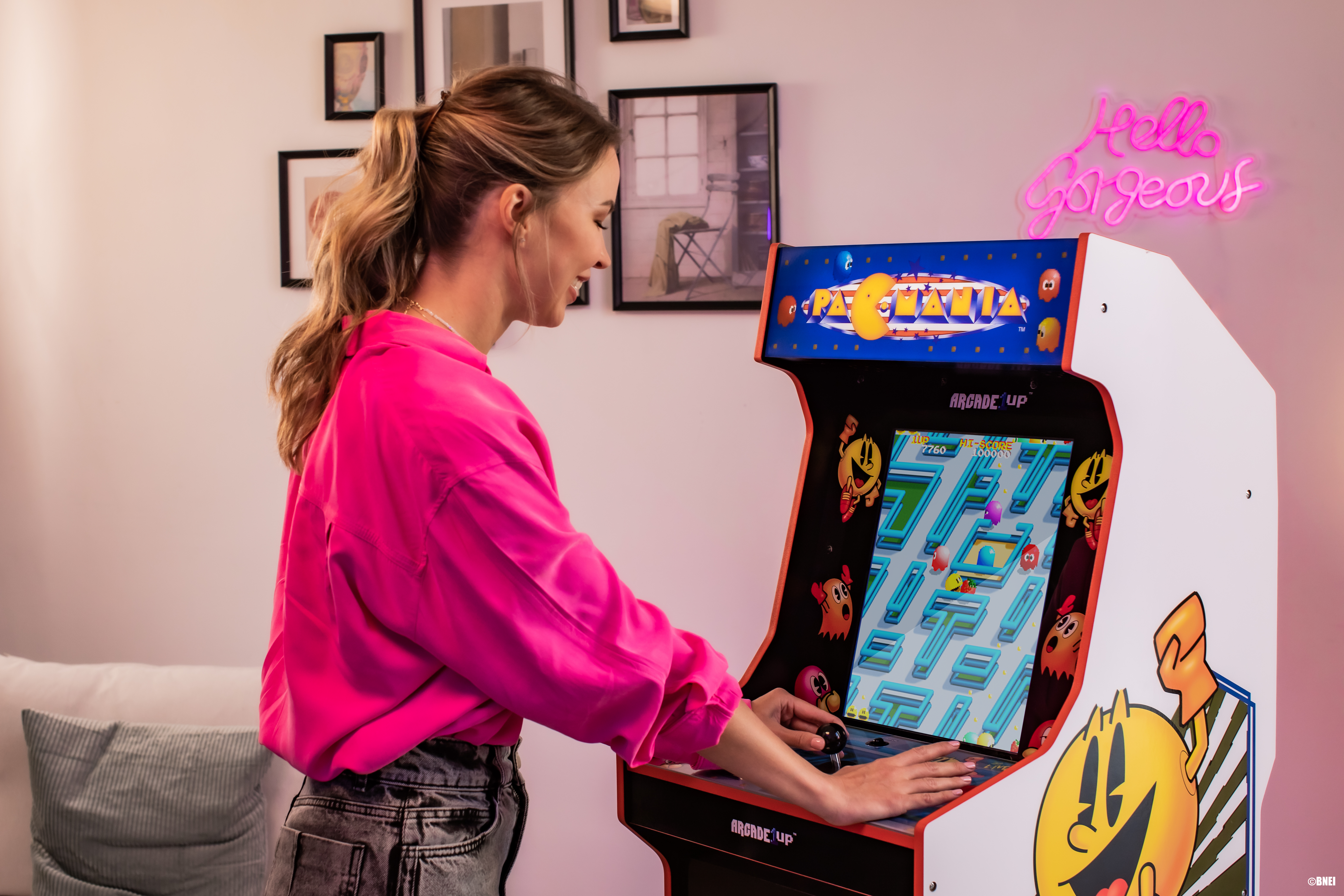 Frau spielt Arcade-Spiel. Das Spiel ist Pac-Man. 'Hello Gorgeous' Neon-Schild im Hintergrund.