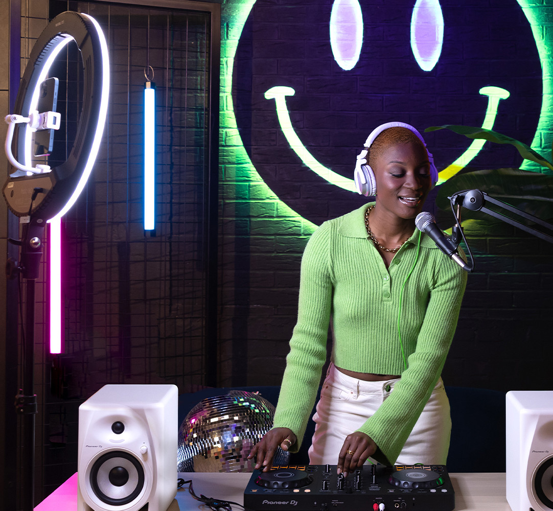 A woman DJing with equipment, a ring light, and a smiley face neon sign.