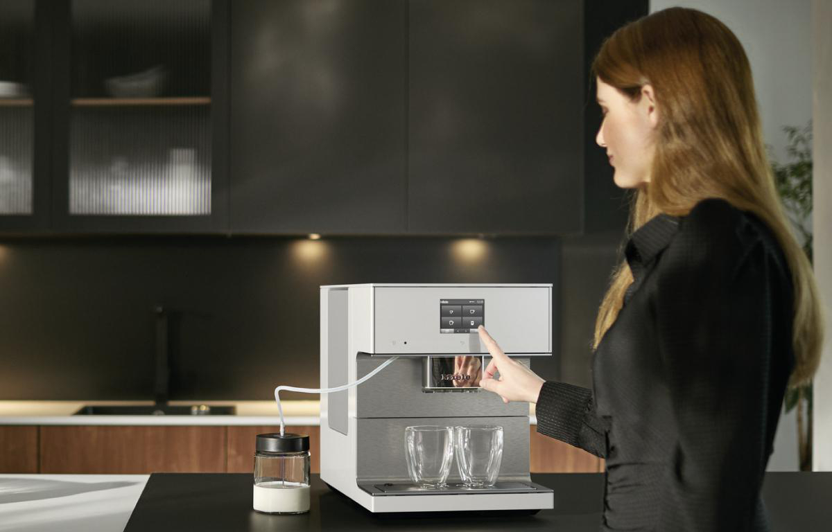 Frau bedient Kaffeemaschine in moderner Küche. Zwei Gläser stehen bereit.
