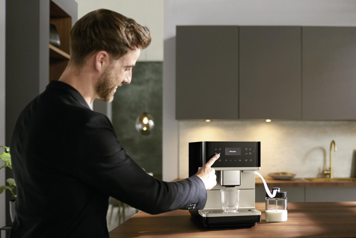 Ein Mann in schwarzer Jacke bedient eine Miele Kaffeemaschine, mit Glas und Milchbehälter.