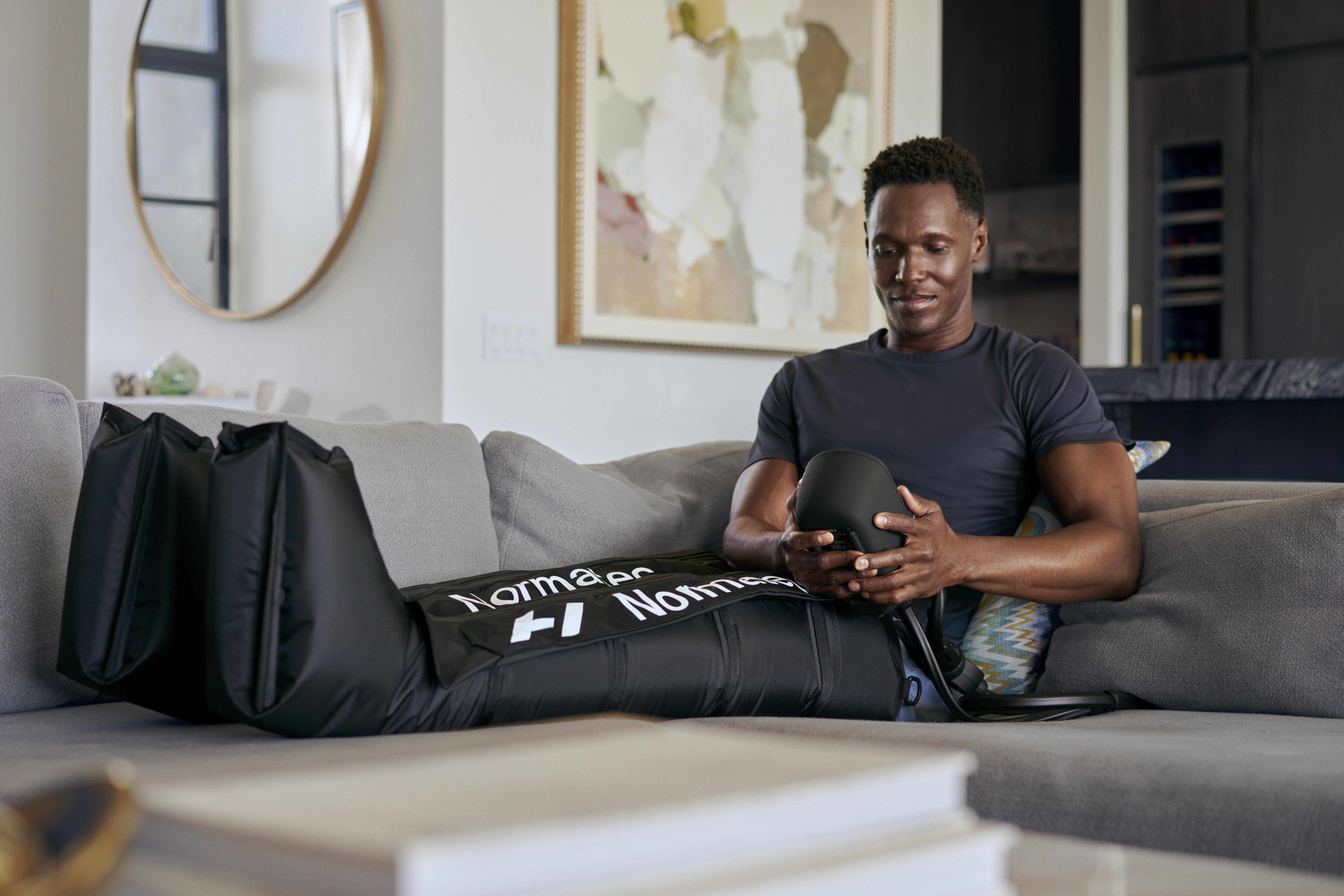 Mann in schwarzem Hemd benutzt Bein-Kompressionsgerät. Sitzt auf grauem Sofa. Umgebung zu Hause.