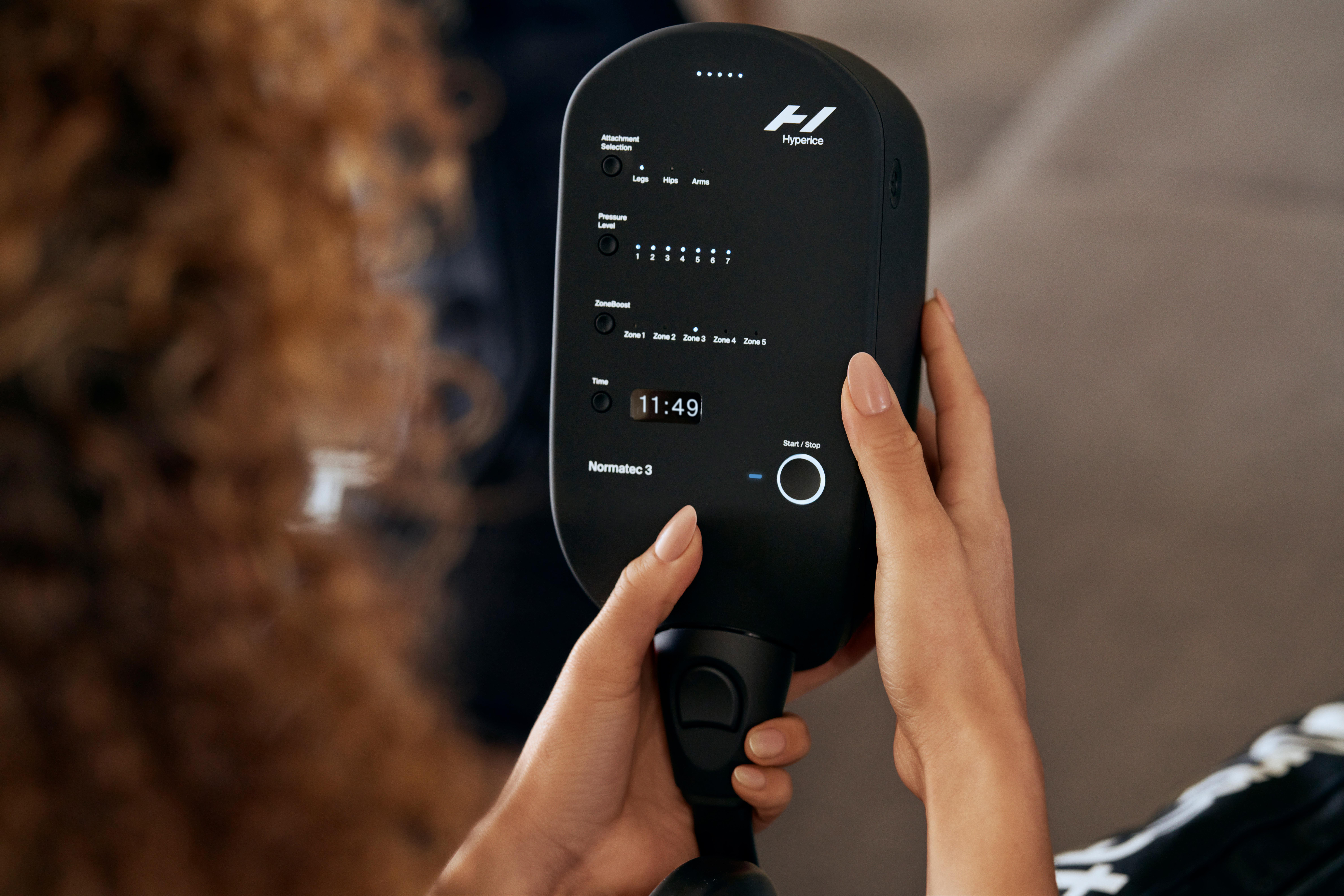 Eine Person hält ein schwarzes Hyperice Normatec 3 Gerät. Es hat Knöpfe und ein Display.