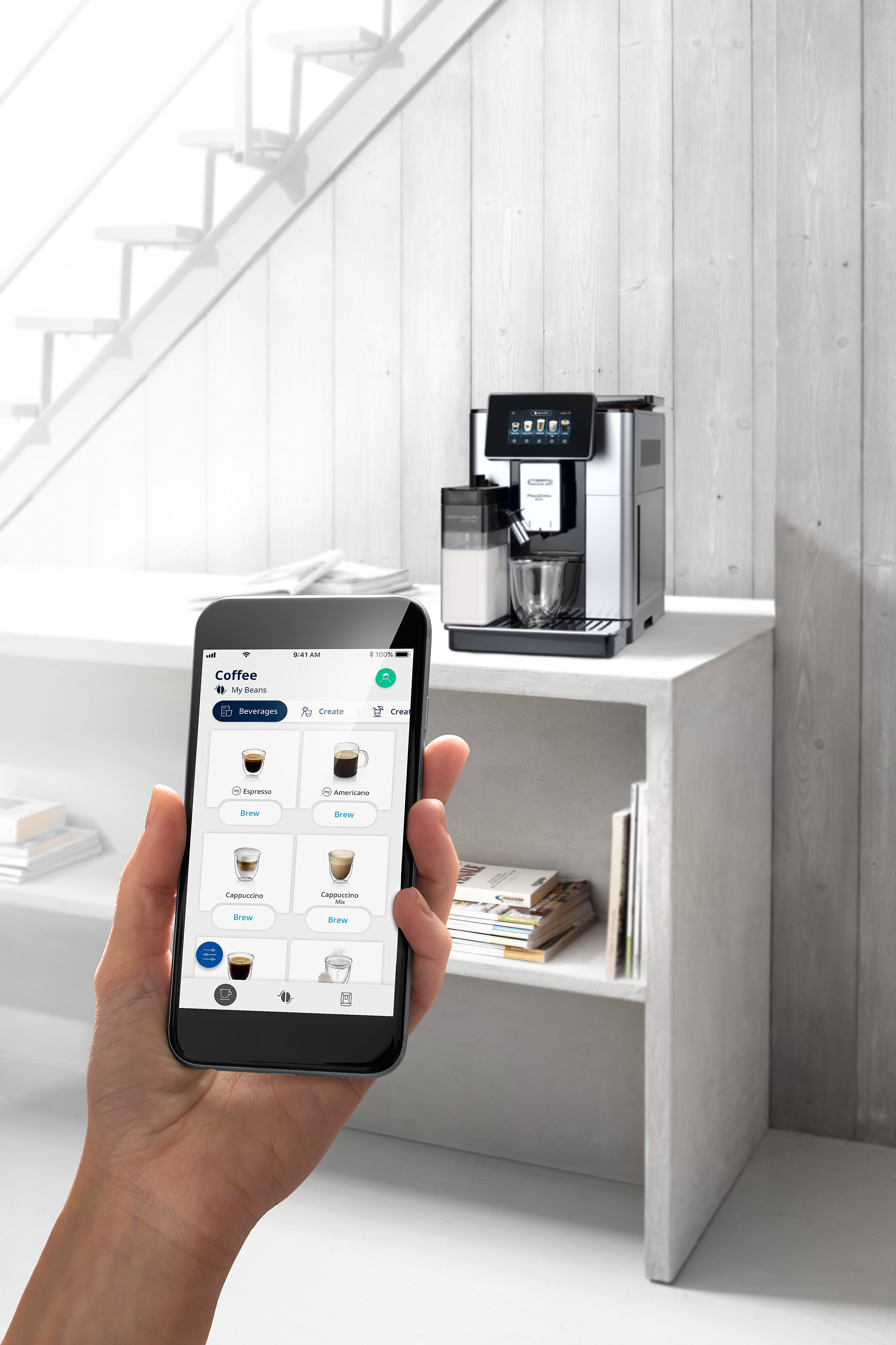Eine Hand hält ein Smartphone mit Kaffeegetränkeoptionen. Im Hintergrund befindet sich eine silberne Kaffeemaschine.