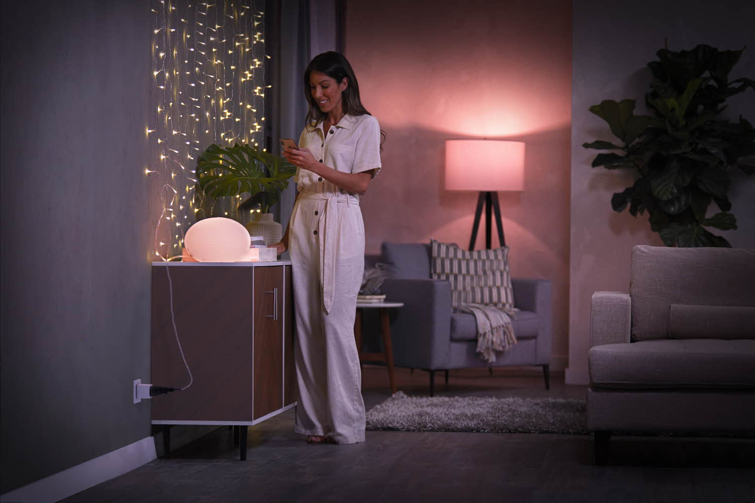 Eine Frau in einem weißen Overall benutzt ein Telefon. Hinter ihr sind eine Lampe und Lichterketten. Im Hintergrund sind eine Couch und Pflanzen.