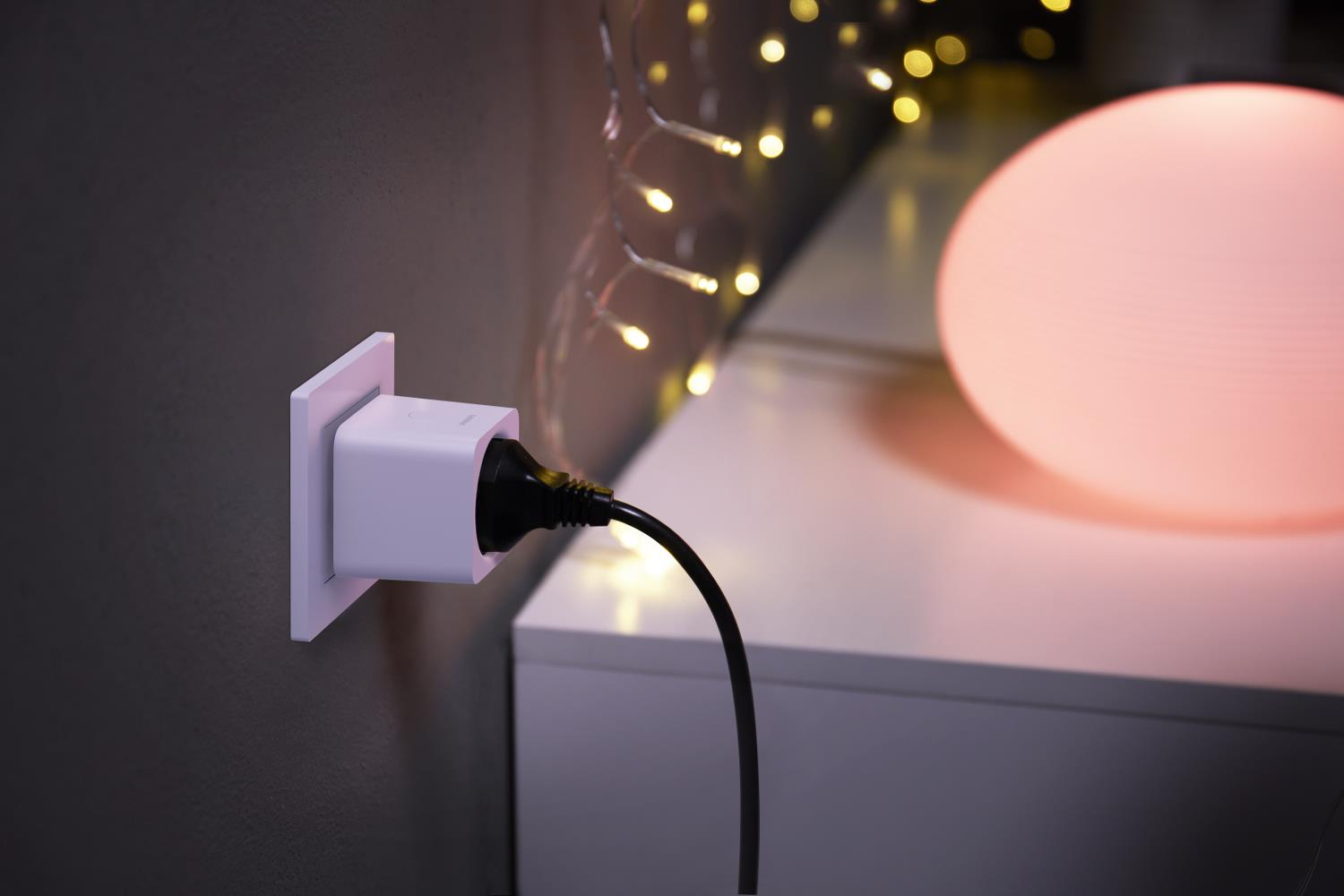 Ein weißer Smart-Stecker ist in eine Steckdose gesteckt. Ein schwarzes Kabel führt zu einem Nachttisch. Eine leuchtende Kugel liegt auf dem Nachtisch.
