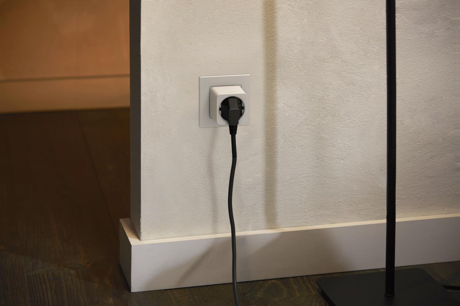 Weiße Wand mit Stecker und schwarzem Kabel. Holzboden im Vordergrund und eine schwarze Lampe rechts.