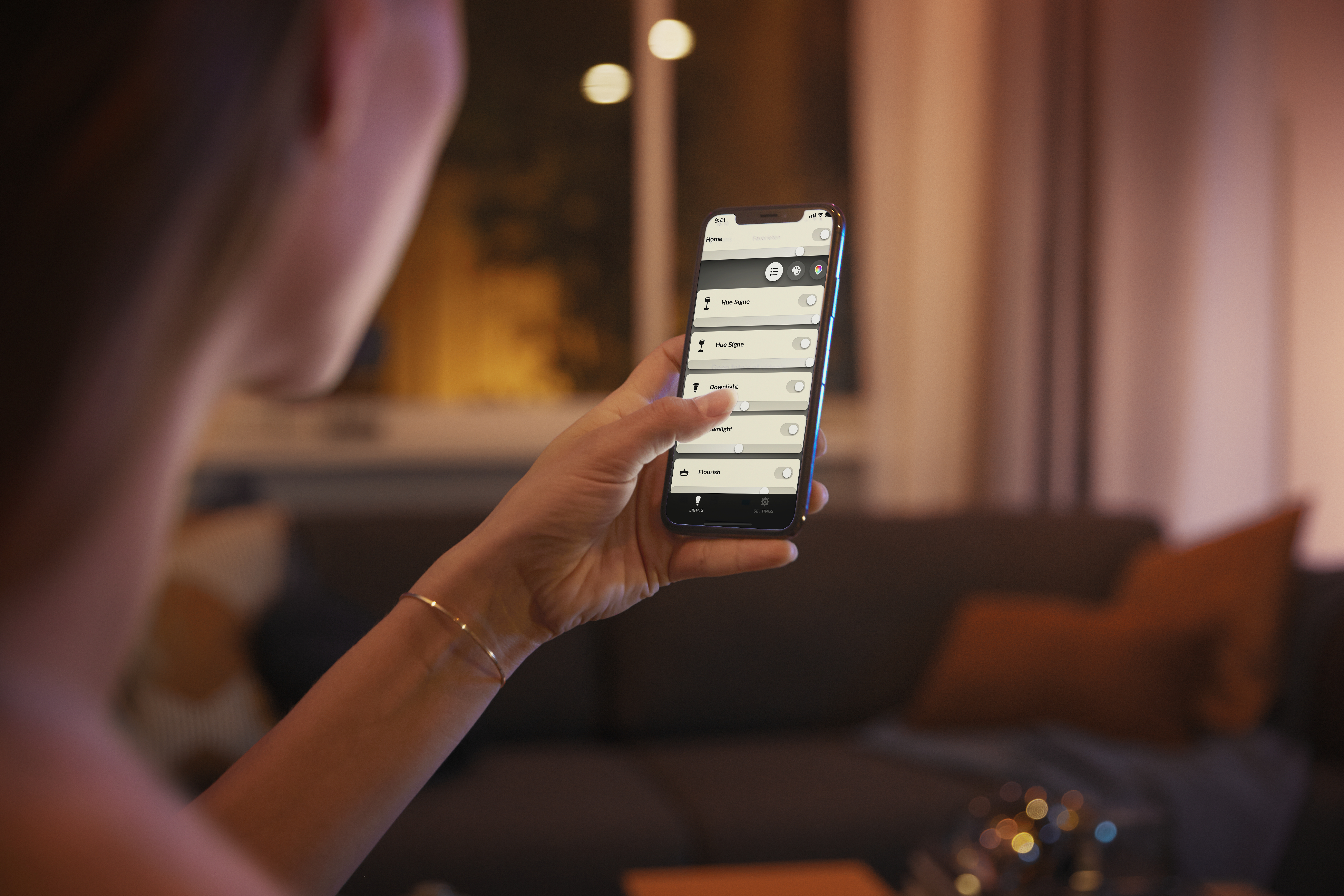 Eine Person hält ein Telefon mit einer Smart-Home-App. Der Hintergrund ist ein Wohnzimmer.
