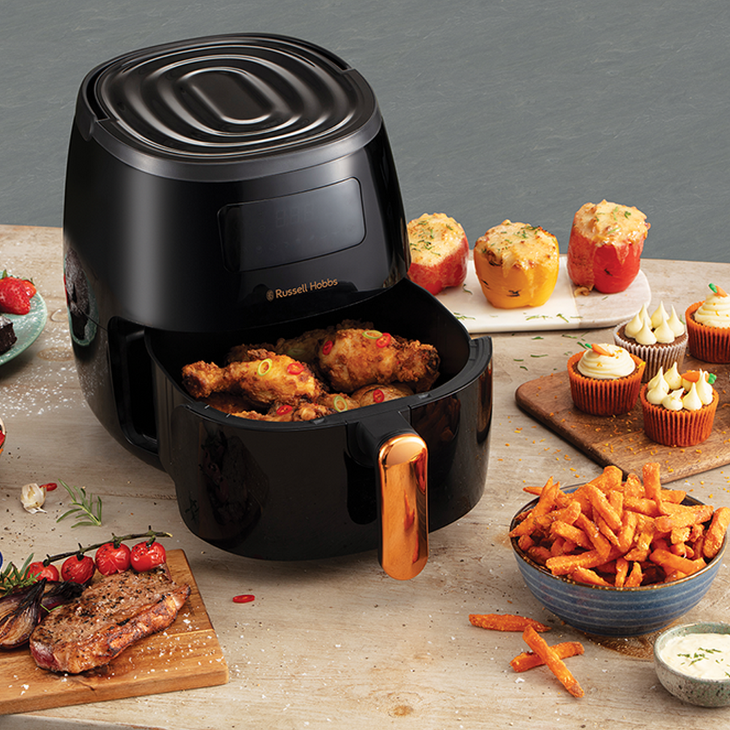 Zwarte airfryer met gefrituurd voedsel en diverse gerechten op houten tafel.