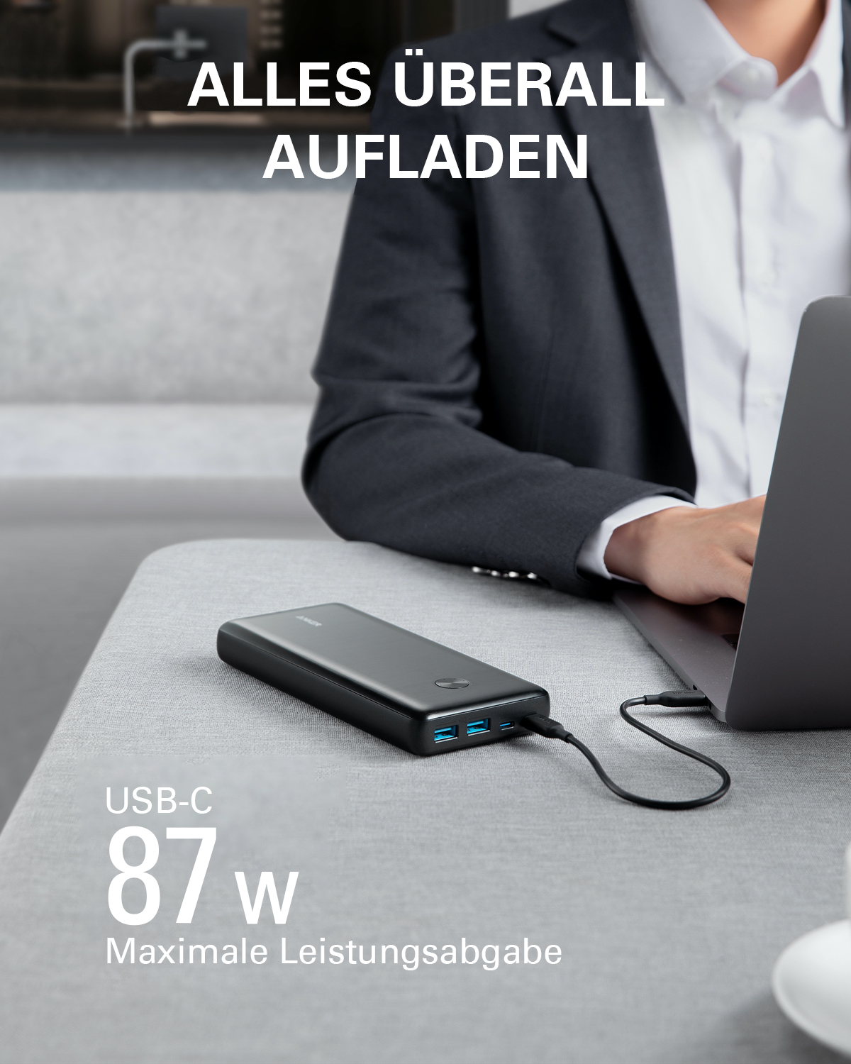 Eine Person benutzt einen Laptop, der an eine schwarze Powerbank auf einem Tisch angeschlossen ist. Der Text lautet 'Alles Überall Aufladen' und 'USB-C 87w'.