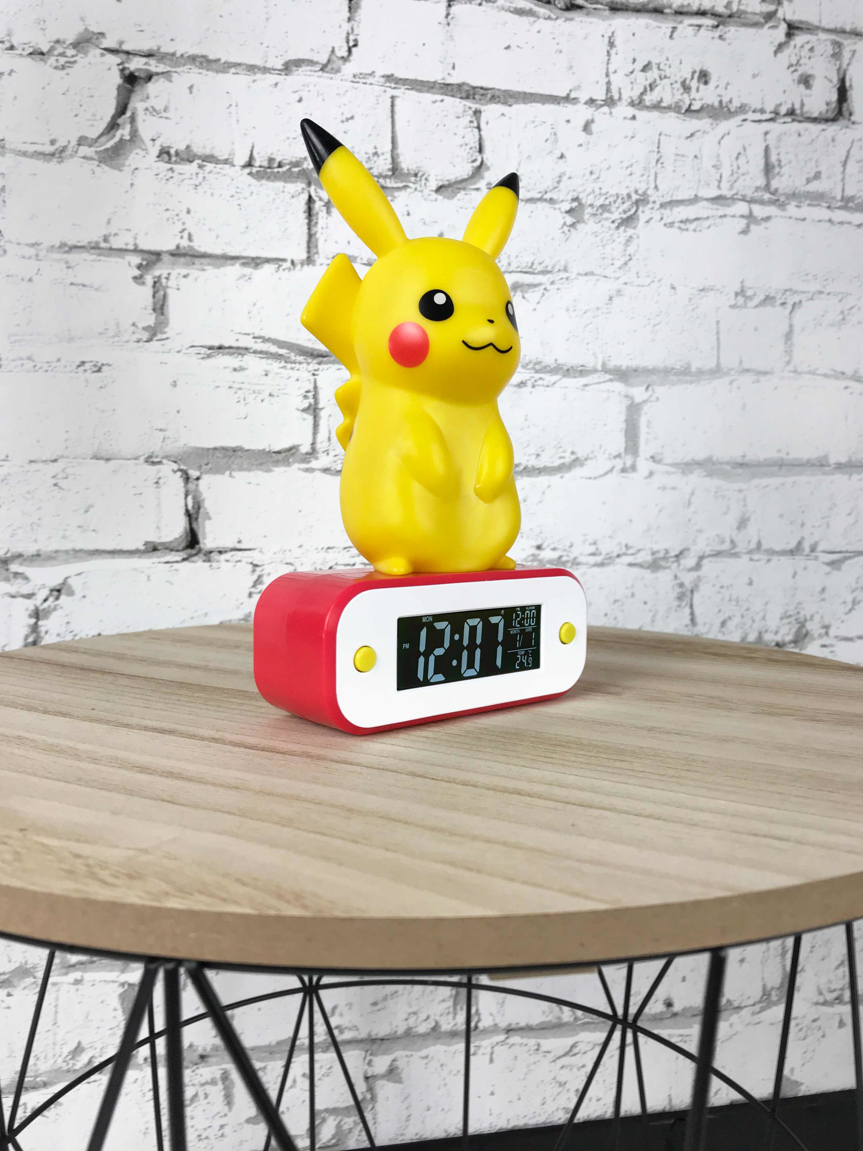 Una sveglia Pikachu gialla, con una base rossa. È su un tavolo di legno davanti a un muro di mattoni.