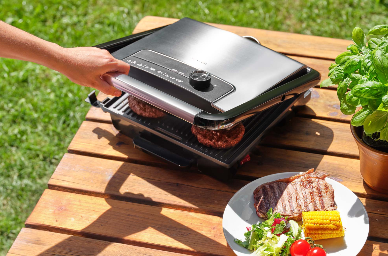 Grill, amin burgerek sülnek. Steak, kukorica és saláta a tányéron. Fa asztal, zöld fű.