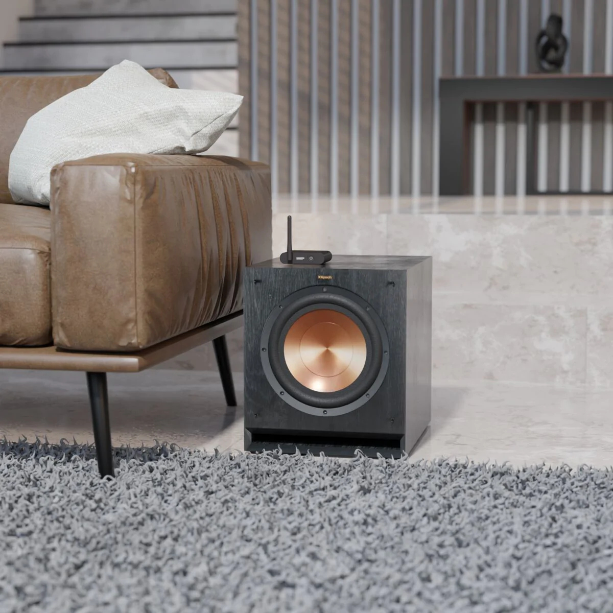 Een zwarte subwoofer met koperkleurige conus staat op een grijs kleed naast een bruine leren bank.