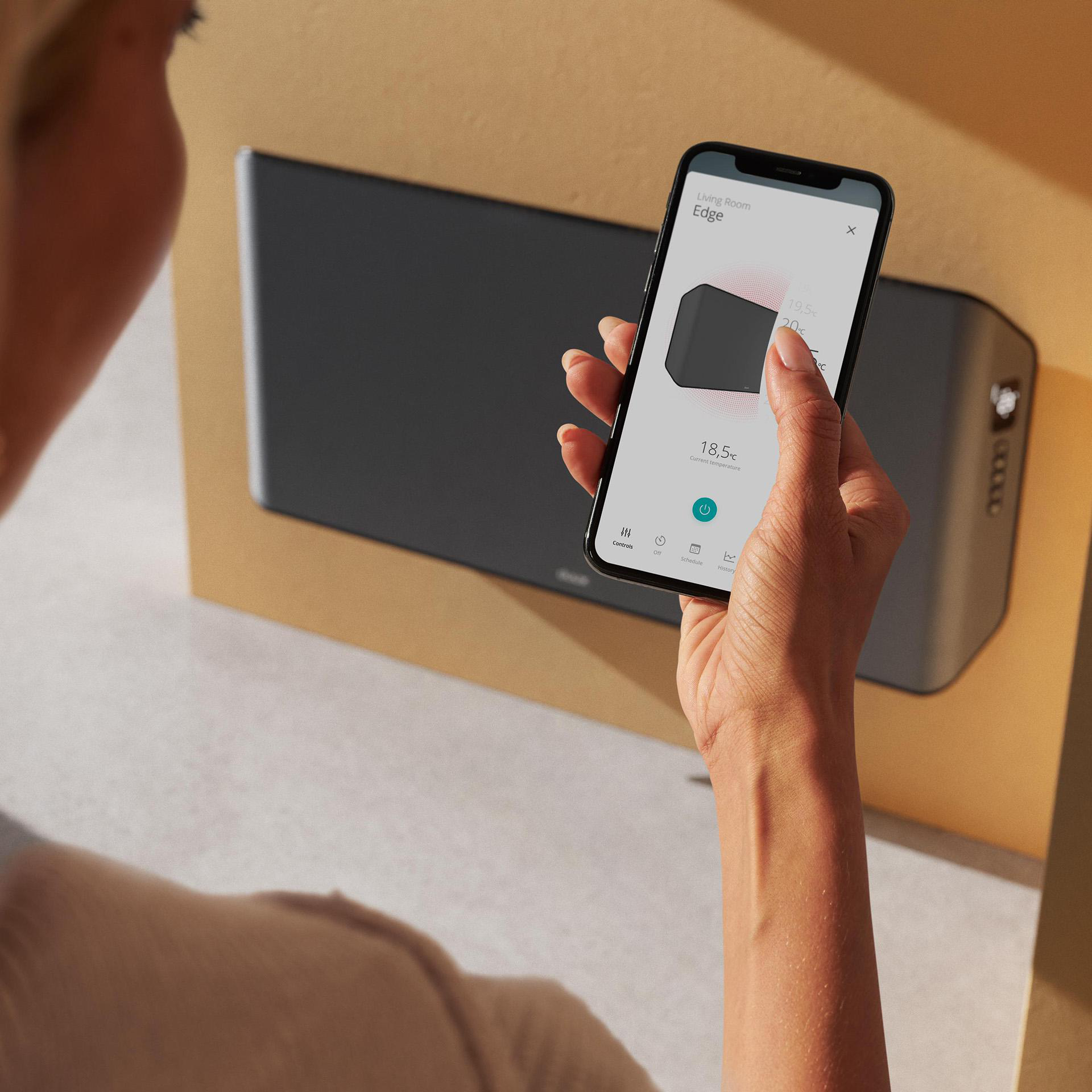 Frau hält ein Telefon mit Smart-Home-Oberfläche in der Nähe eines an der Wand montierten Geräts.