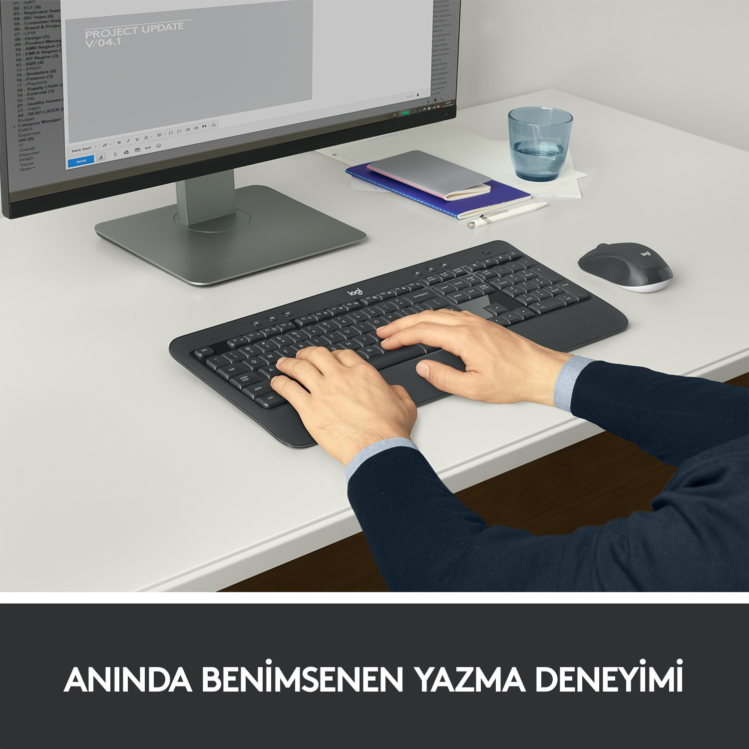 Beyaz bir masada siyah bir klavyede yazan eller. Bir monitör, fare ve bir bardak su.