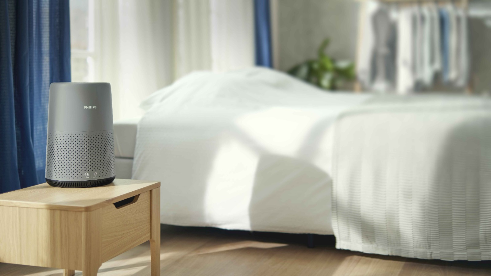 Ahşap bir komodin üzerinde bir Philips hava temizleyici. Arka planda mavi perdelerle bir yatak ve pencere.