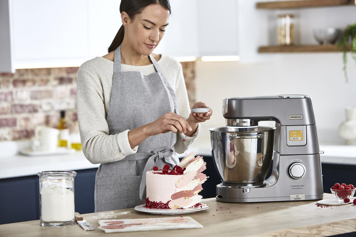 Frau dekoriert Kuchen, Mixer im Hintergrund. Küchenumgebung mit Mehl und Himbeeren.