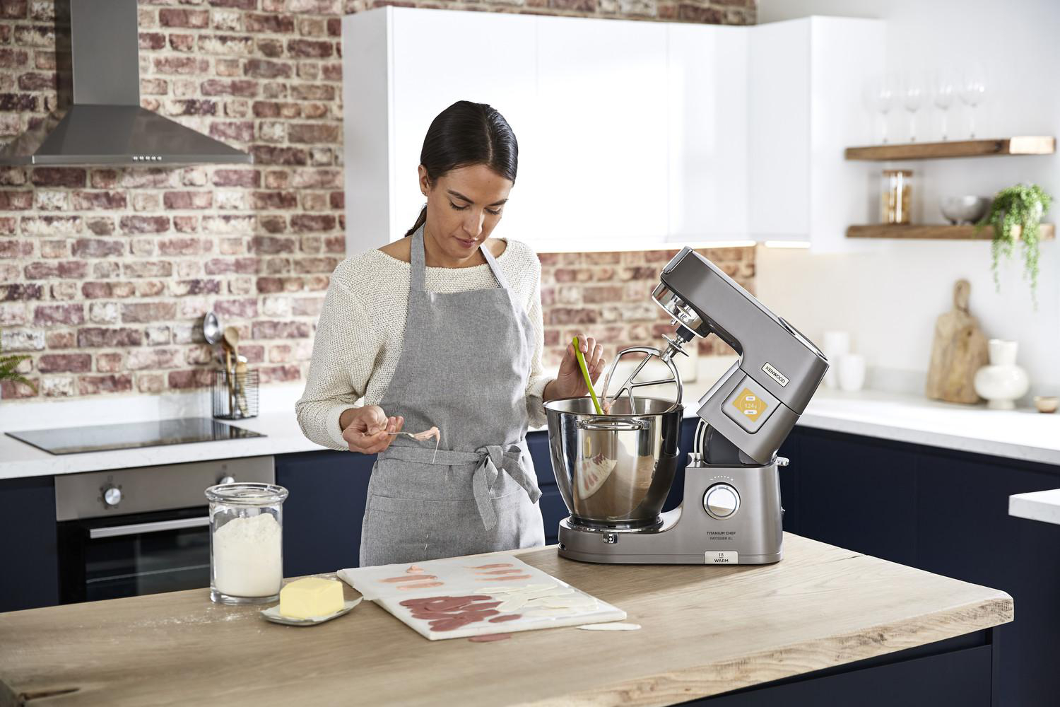 Eine Frau mit Schürze benutzt einen Standmixer in einer Küche mit Backsteinwänden und Holz-Arbeitsplatten.