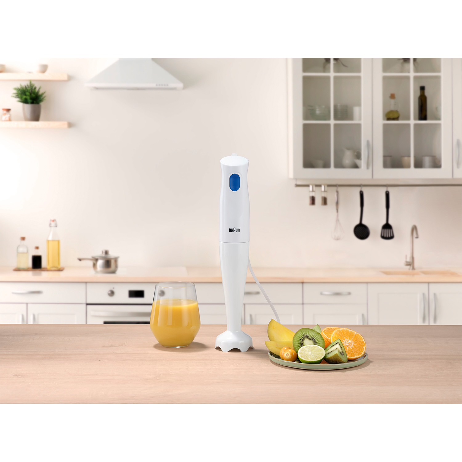 Frullatore a immersione bianco con pulsante blu, succo d'arancia e frutta sul bancone.