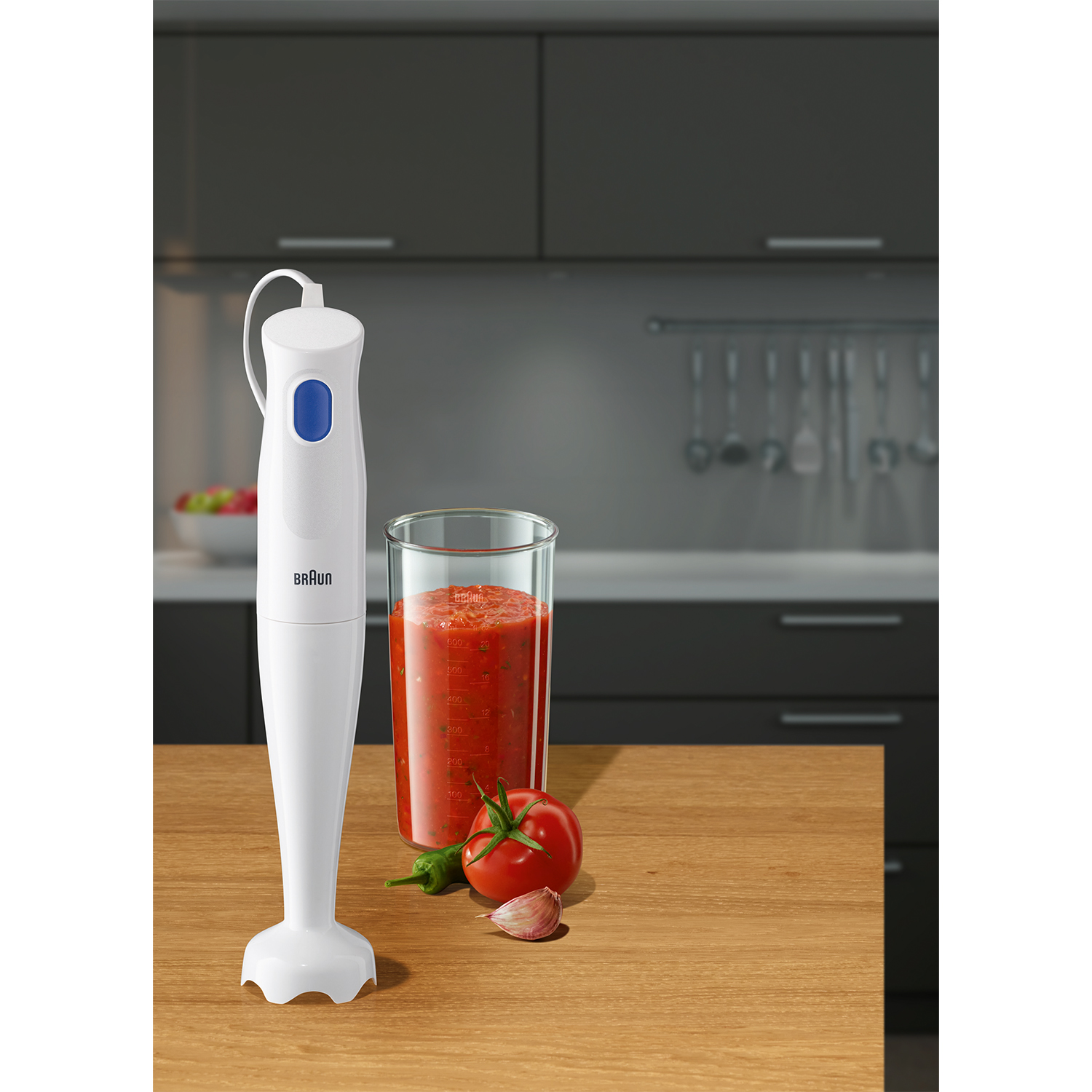 Frullatore a immersione bianco con pulsante blu. Un bicchiere di salsa di pomodoro, pomodoro e aglio nelle vicinanze.