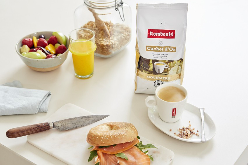 Scène de petit-déjeuner : café, bagel, coupe de fruits, jus et céréales, sur une table blanche.