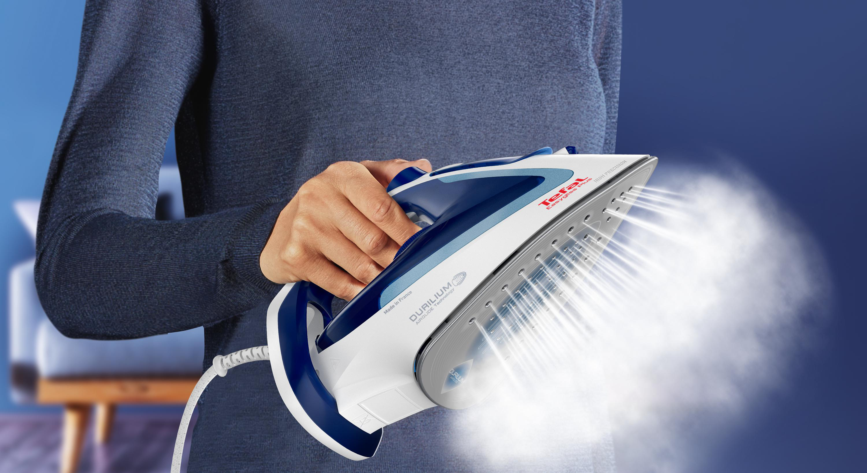 Eine Person hält ein Tefal-Bügeleisen, das dampft. Das Bügeleisen ist blau und weiß. Der Hintergrund ist eine blaue Wand.