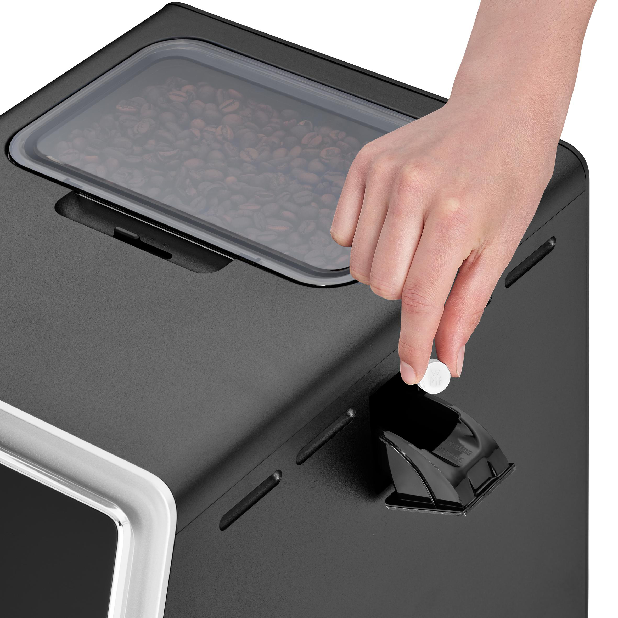 Hand, die eine Tablette in das Bohnenfach einer schwarzen Kaffeemaschine gibt.
