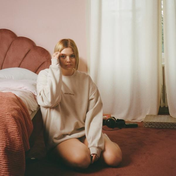 Femme blonde assise sur un lit rose, portant un sweat-shirt, regardant vers le bas, dans une pièce rose.