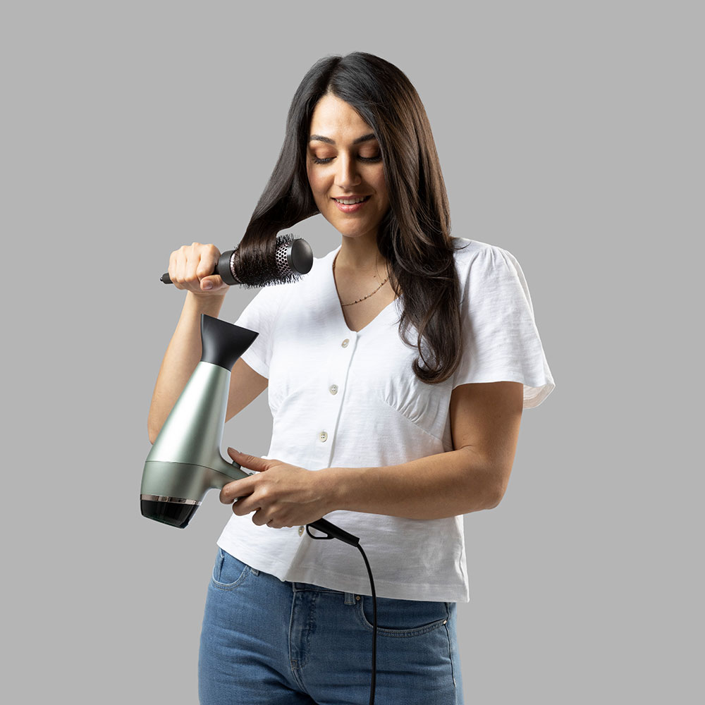 Donna che usa un asciugacapelli e una spazzola rotonda, acconciandosi i capelli. Top bianco e jeans blu.