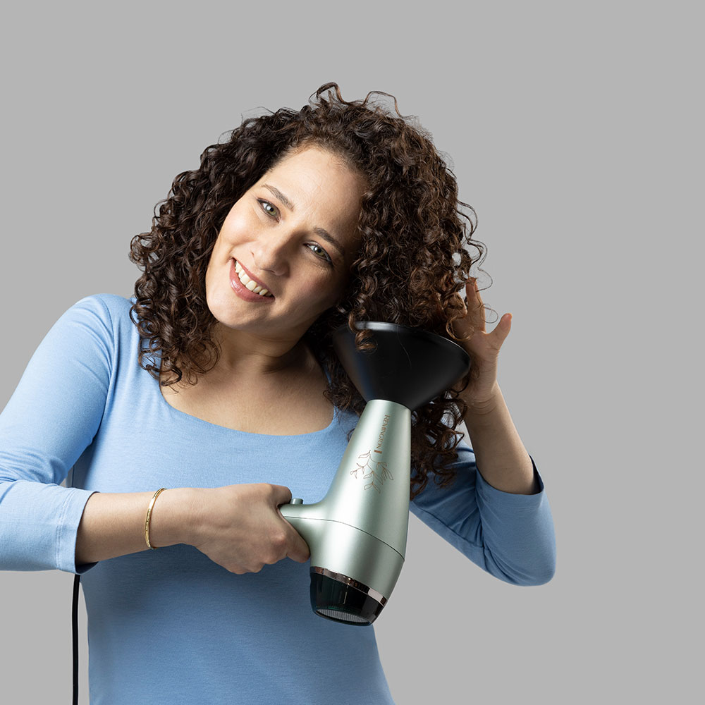 Donna che si asciuga i capelli ricci con un asciugacapelli verde e nero, sorridendo alla fotocamera.