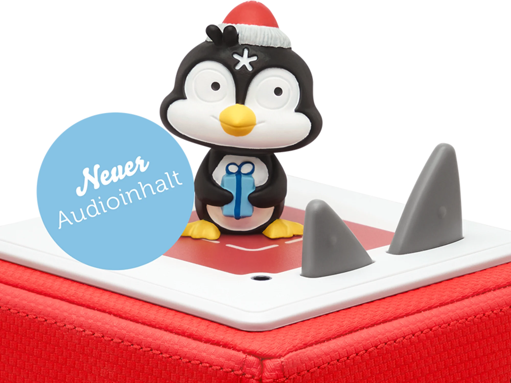 Un giocattolo a forma di pinguino con cappello da Babbo Natale, un regalo e figure simili a squali su una superficie rossa.