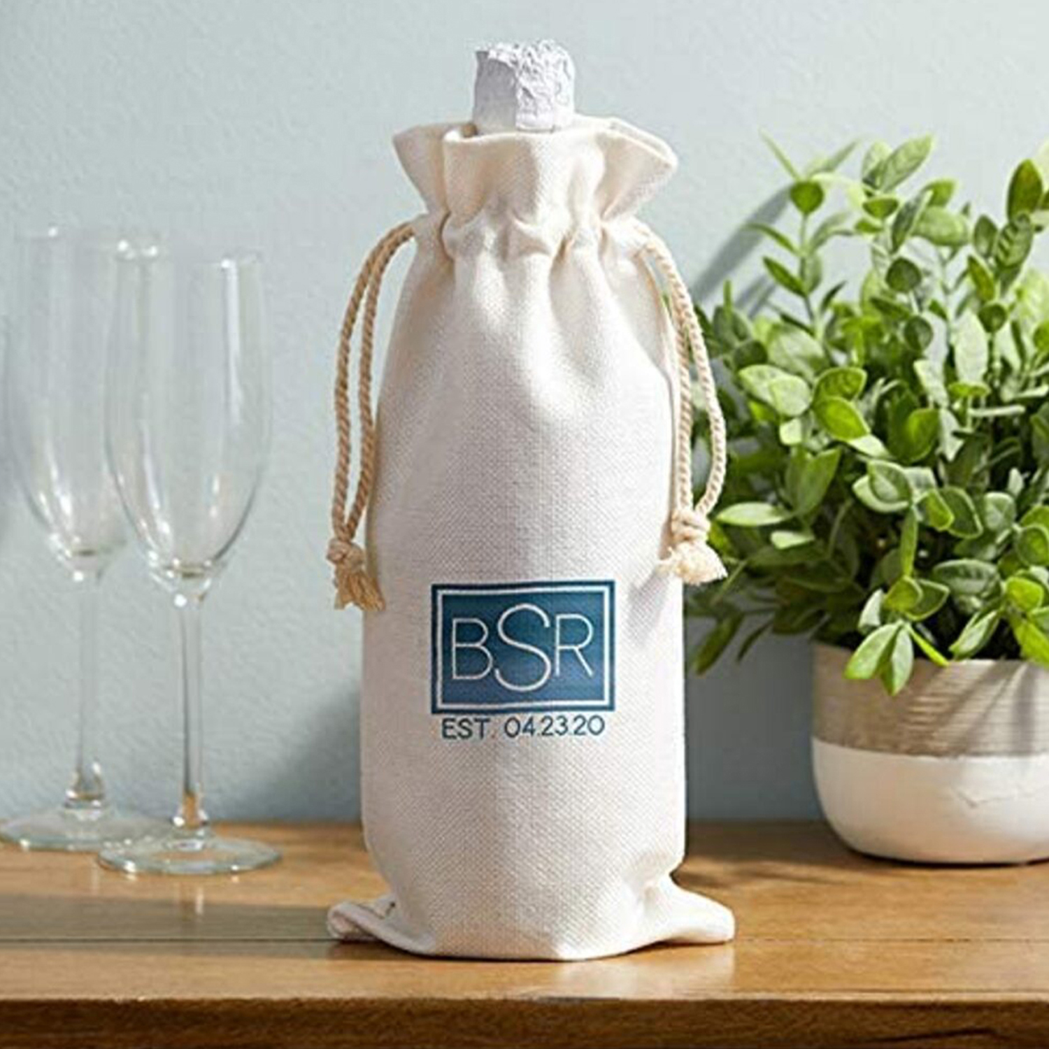 Bottiglia di vino in un sacco bianco con logo blu e cordino. Due bicchieri sullo sfondo.