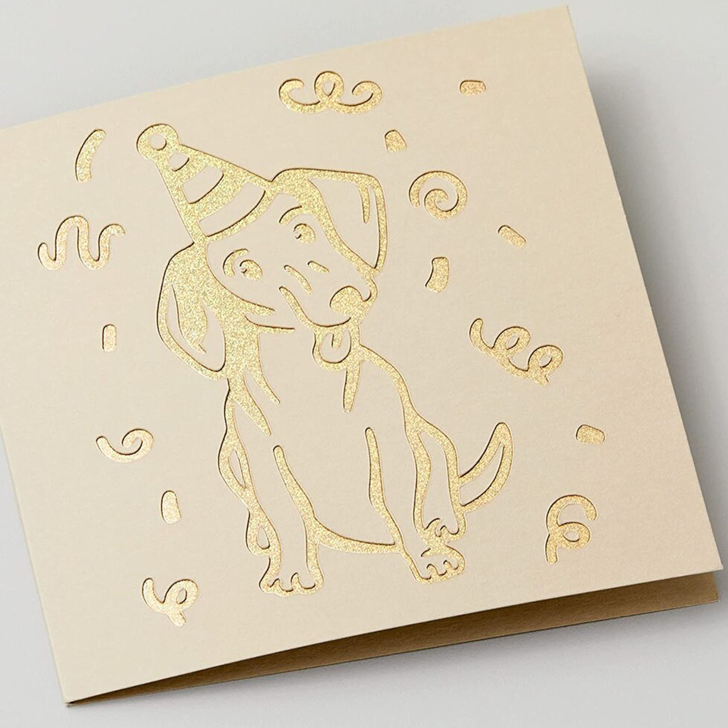 Un biglietto di compleanno con un cane dorato con un cappello da festa. Il disegno è ritagliato dalla carta.