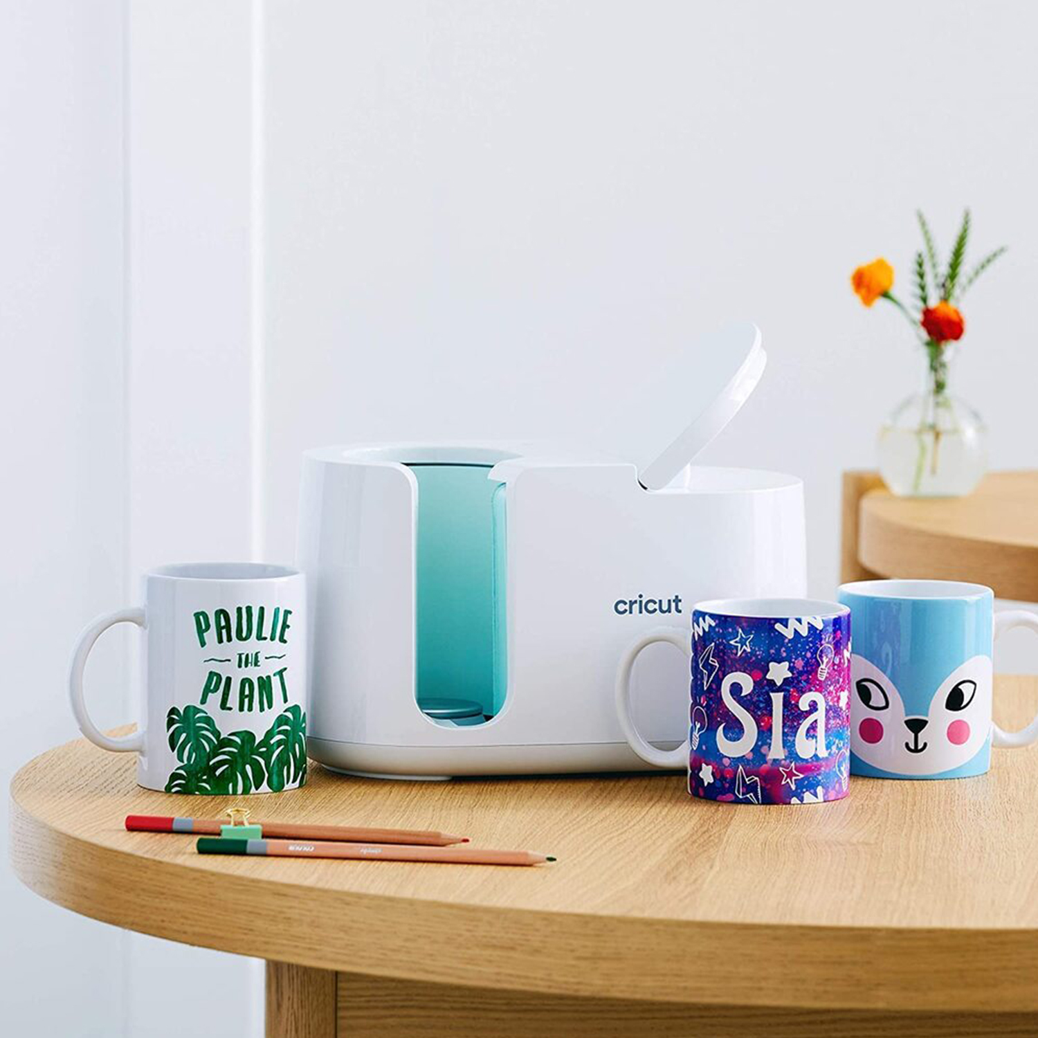 Tazze con disegni su tavolo di legno, con macchina pressa per tazze Cricut.