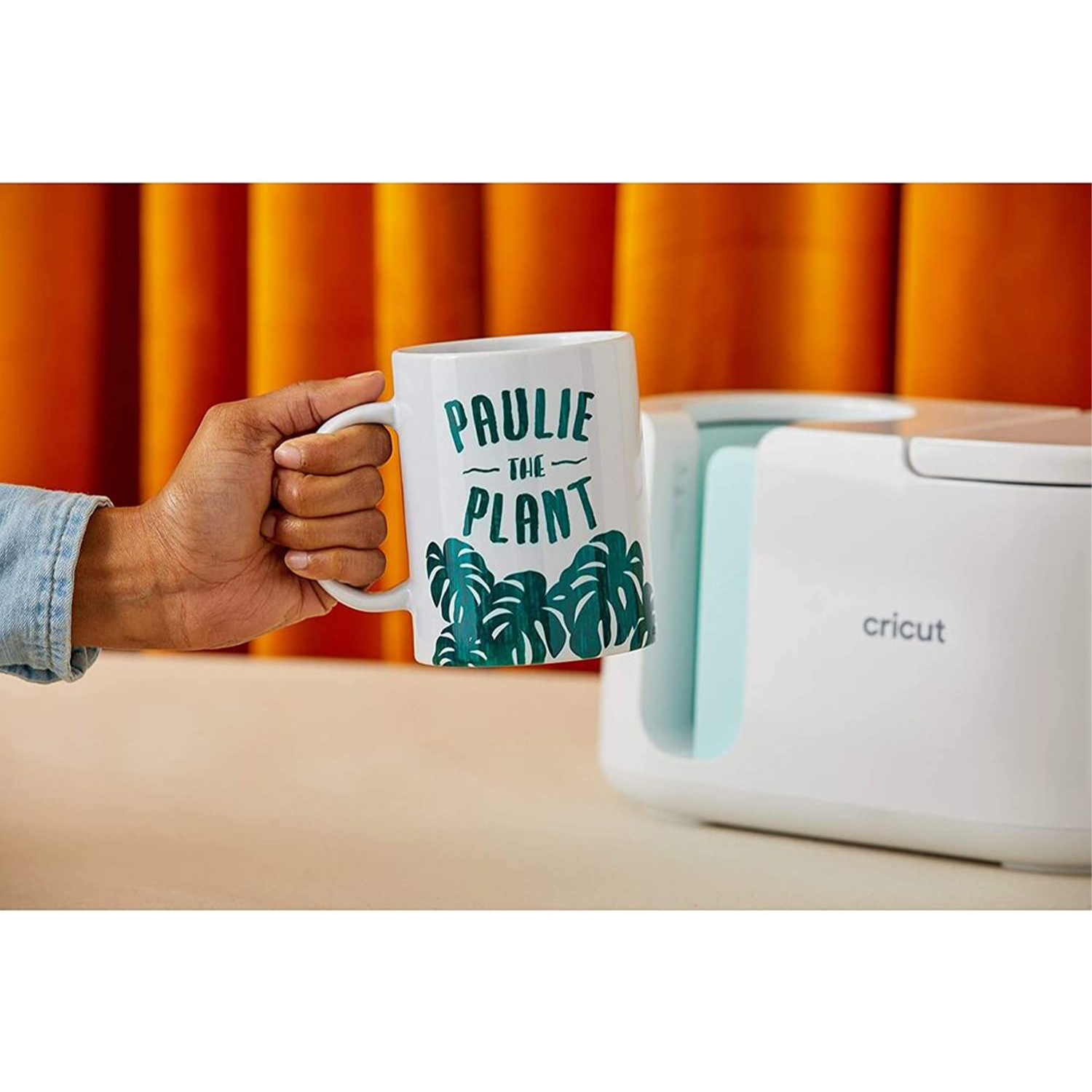 Una persona tiene in mano una tazza con scritto 'Paulie the Plant' vicino a una macchina Cricut.