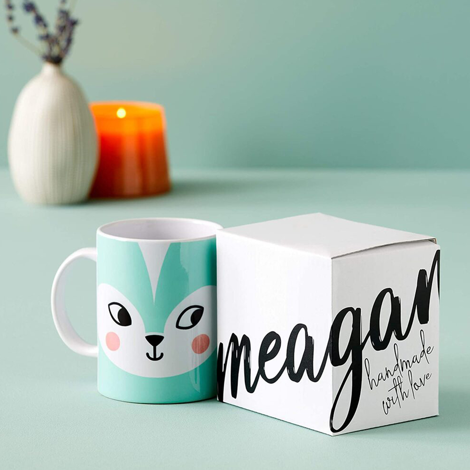 Una tazza turchese con un disegno di volpe e una scatola bianca. Un vaso e una candela sullo sfondo.