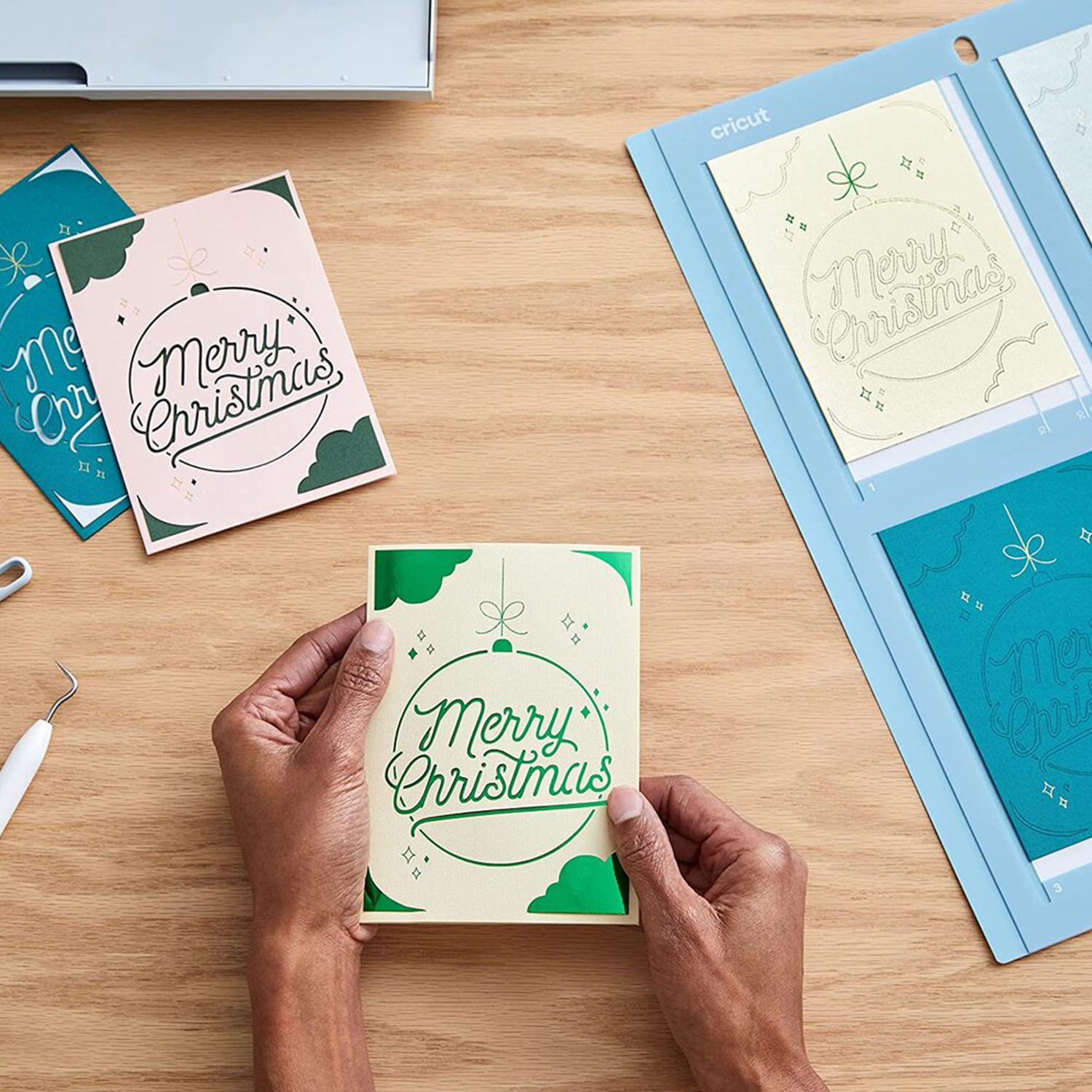 Mani che tengono un biglietto di Natale, con altri biglietti e una macchina Cricut sullo sfondo.