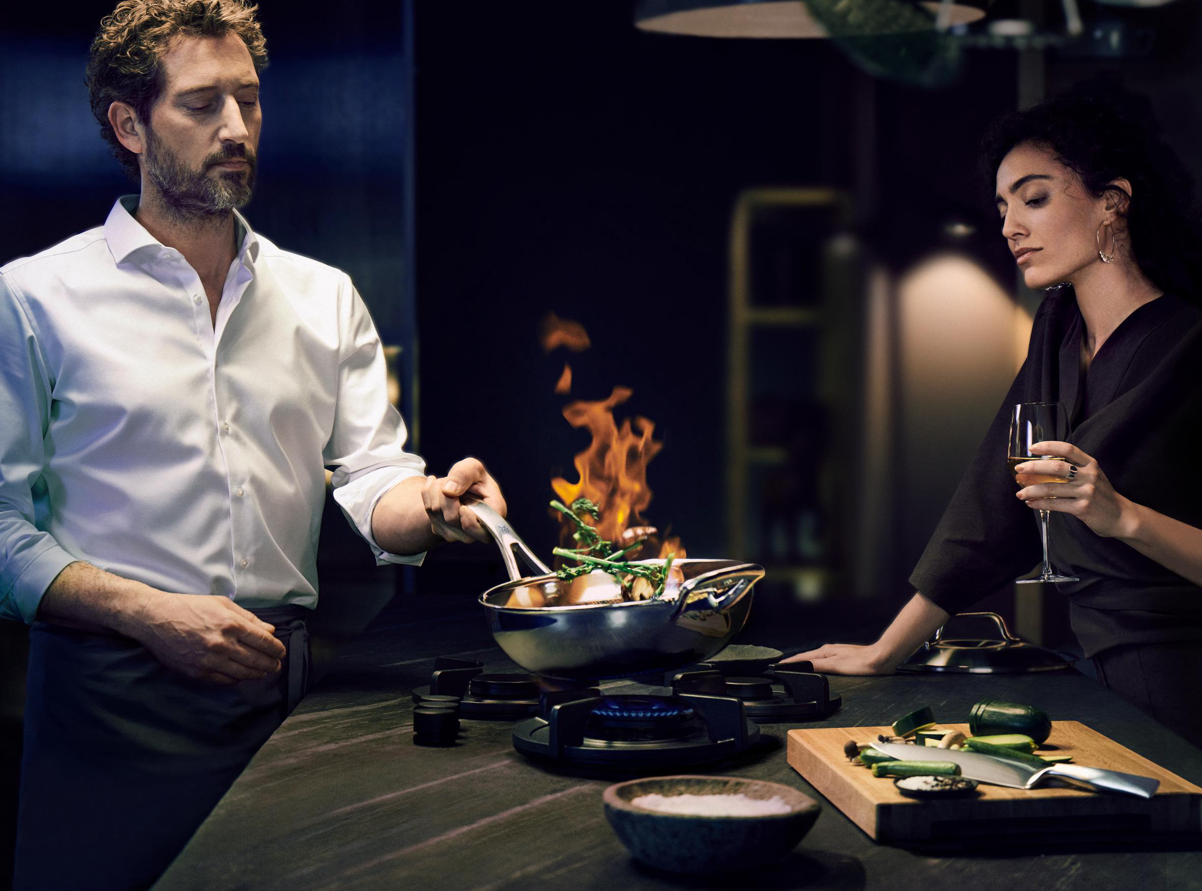 Ein Mann kocht in einer Pfanne mit Flammen, eine Frau mit Wein, eine Küchenumgebung.