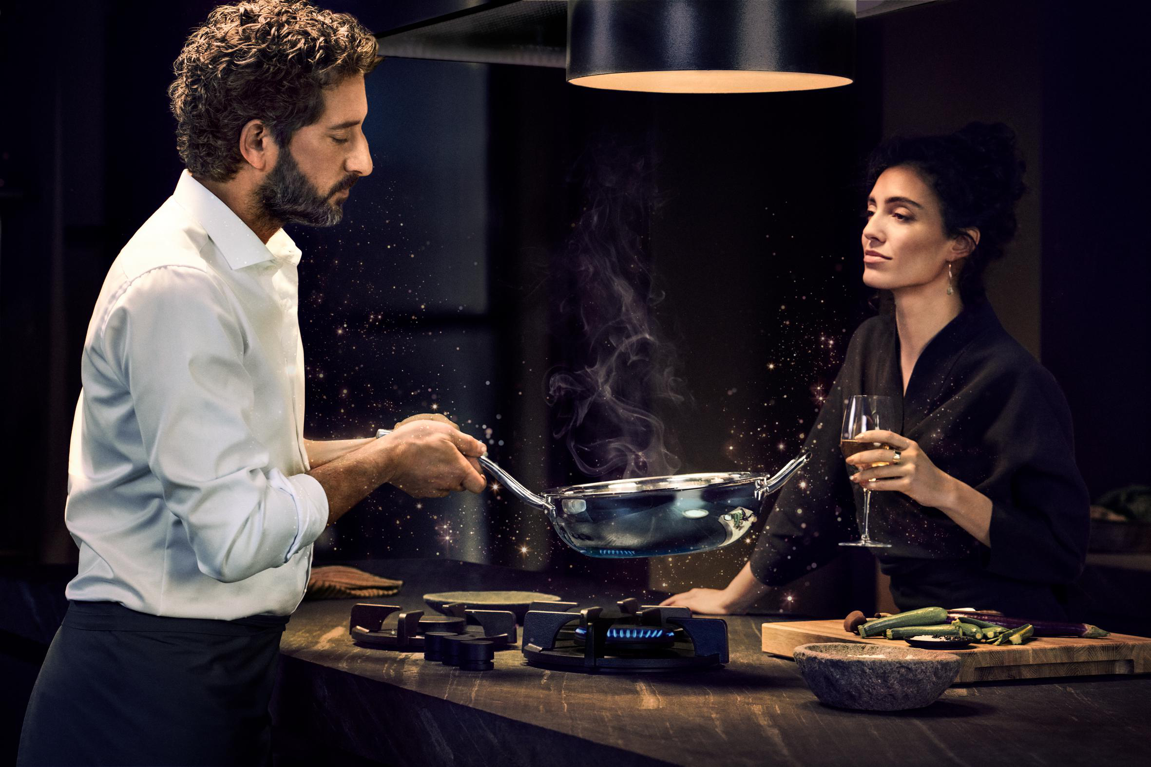Mann kocht in der Küche, Frau hält Glas. Moderne Küche, schwaches Licht, Kochen und Wein trinken.