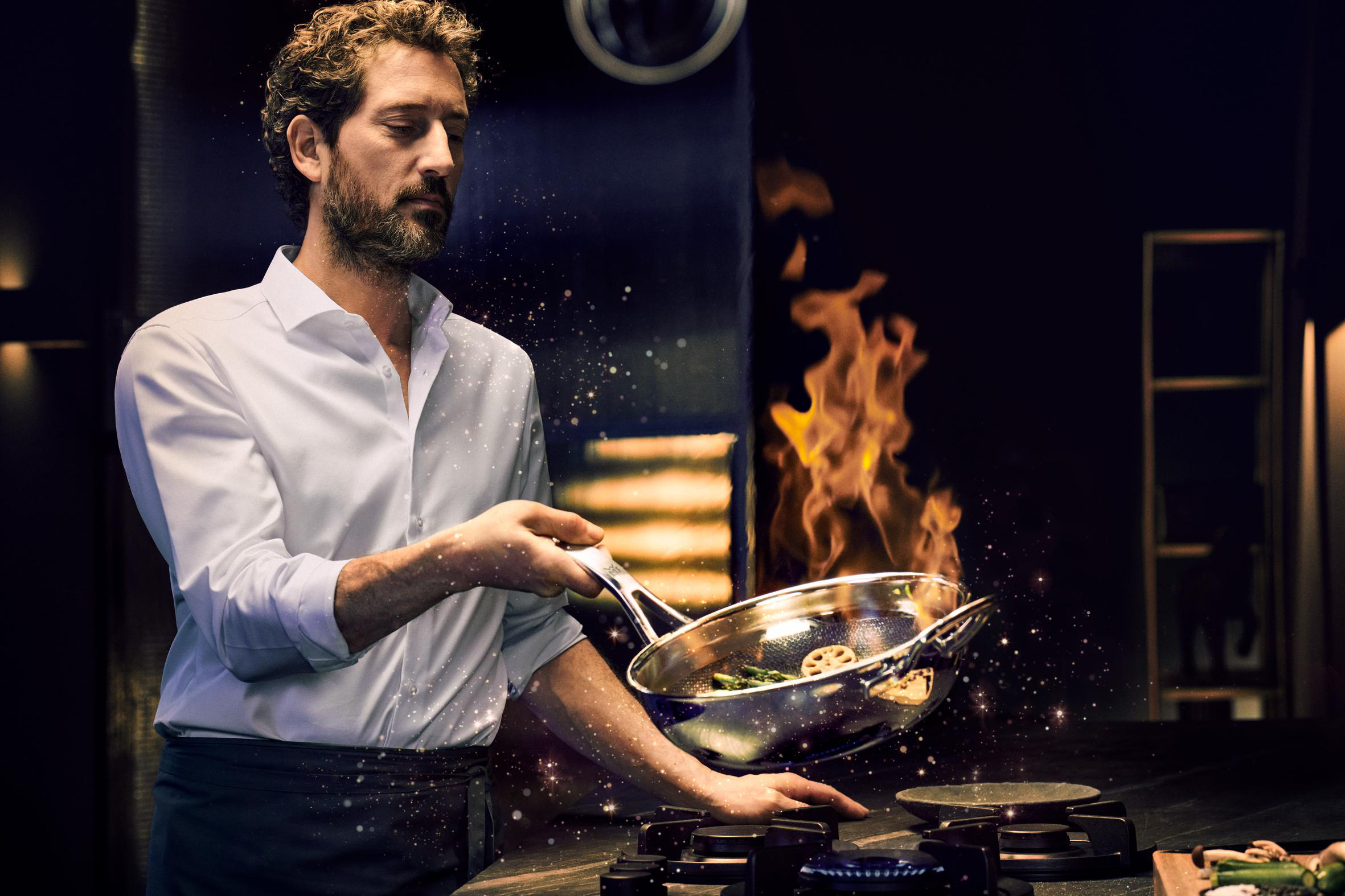 Ein Koch wirft Essen in einer Pfanne über einem Herd mit Flammen. Funken und ein dunkler Küchenhintergrund sind vorhanden.
