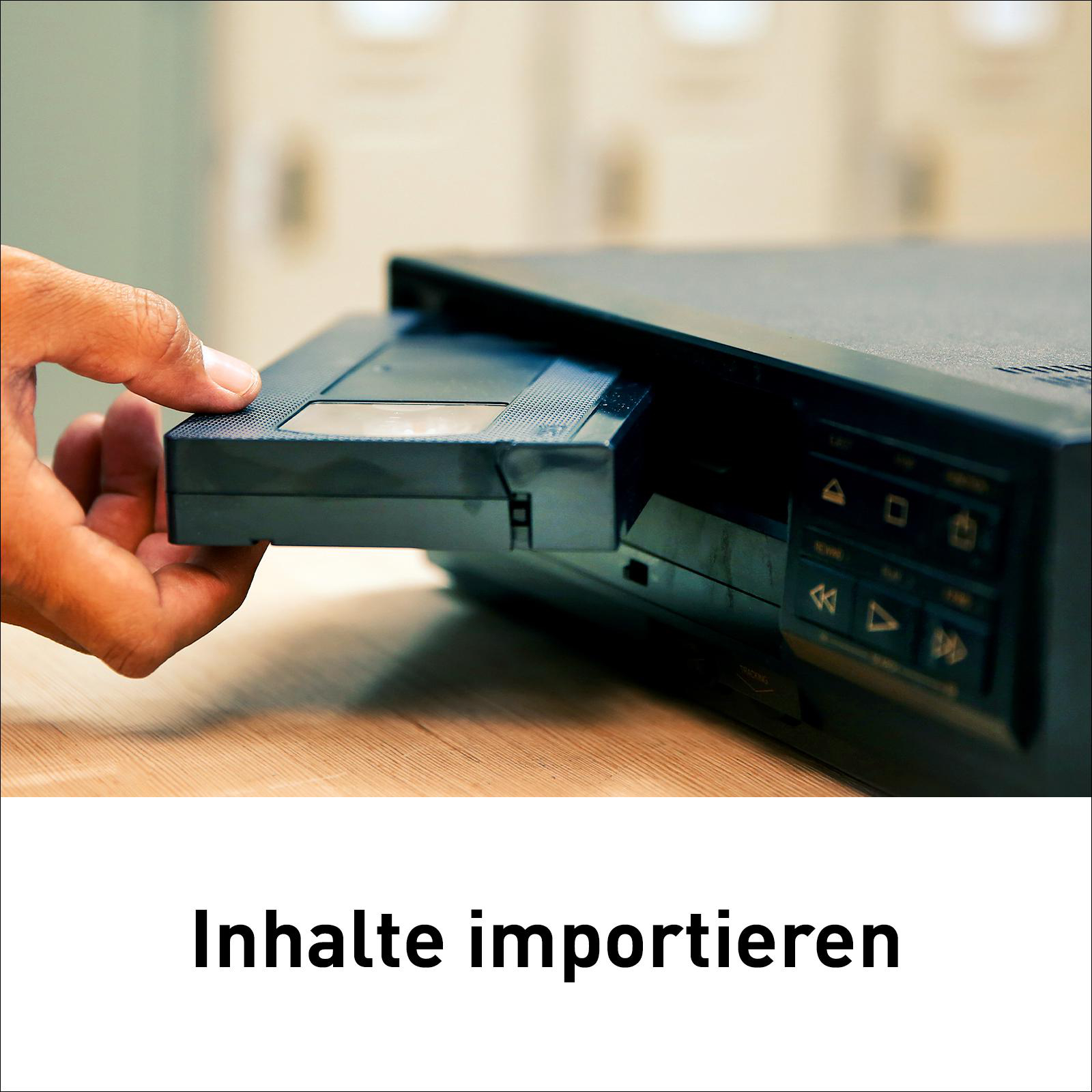 Eine Hand führt eine VHS-Kassette in einen schwarzen Videorekorder auf einem Holztisch ein.