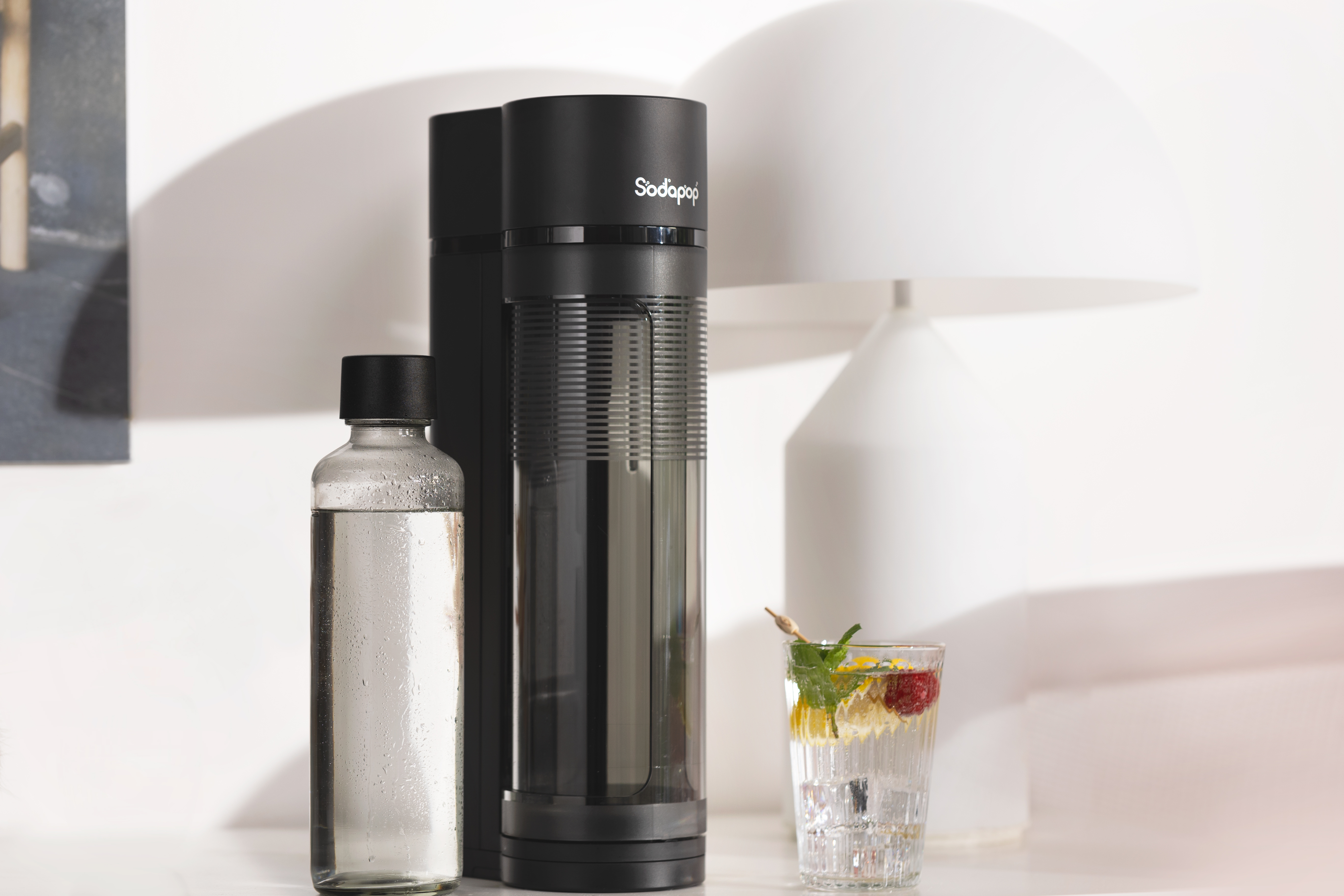 Schwarzer Soda-Maker, klare Flasche und ein Glas mit Eis und Getränk. Weißer Tisch und Lampe.