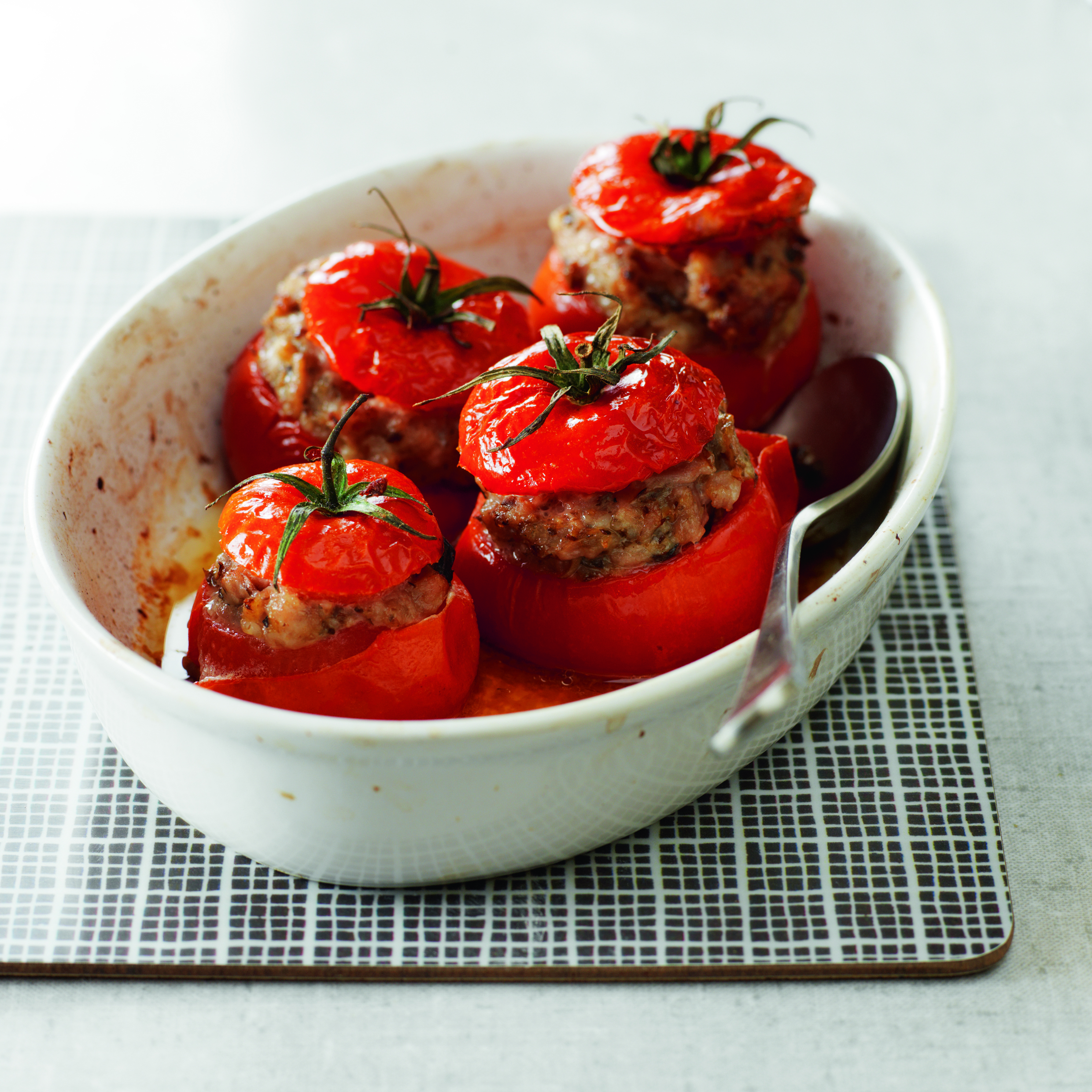 Cuatro tomates rellenos en un plato ovalado blanco con una cuchara sobre una esterilla a cuadros.