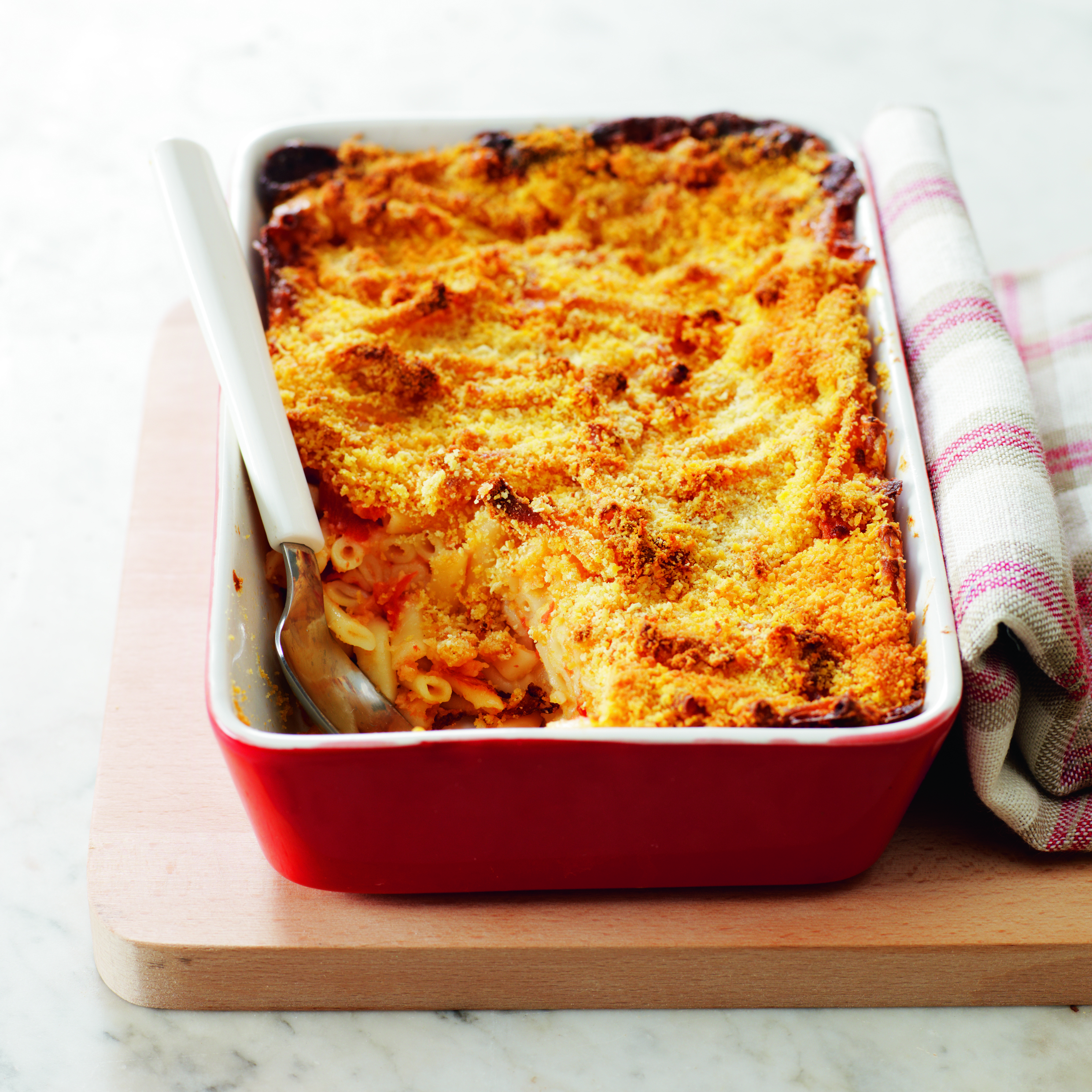 Cazuela de pasta al horno en plato rojo con cuchara sobre tabla de madera y tela.