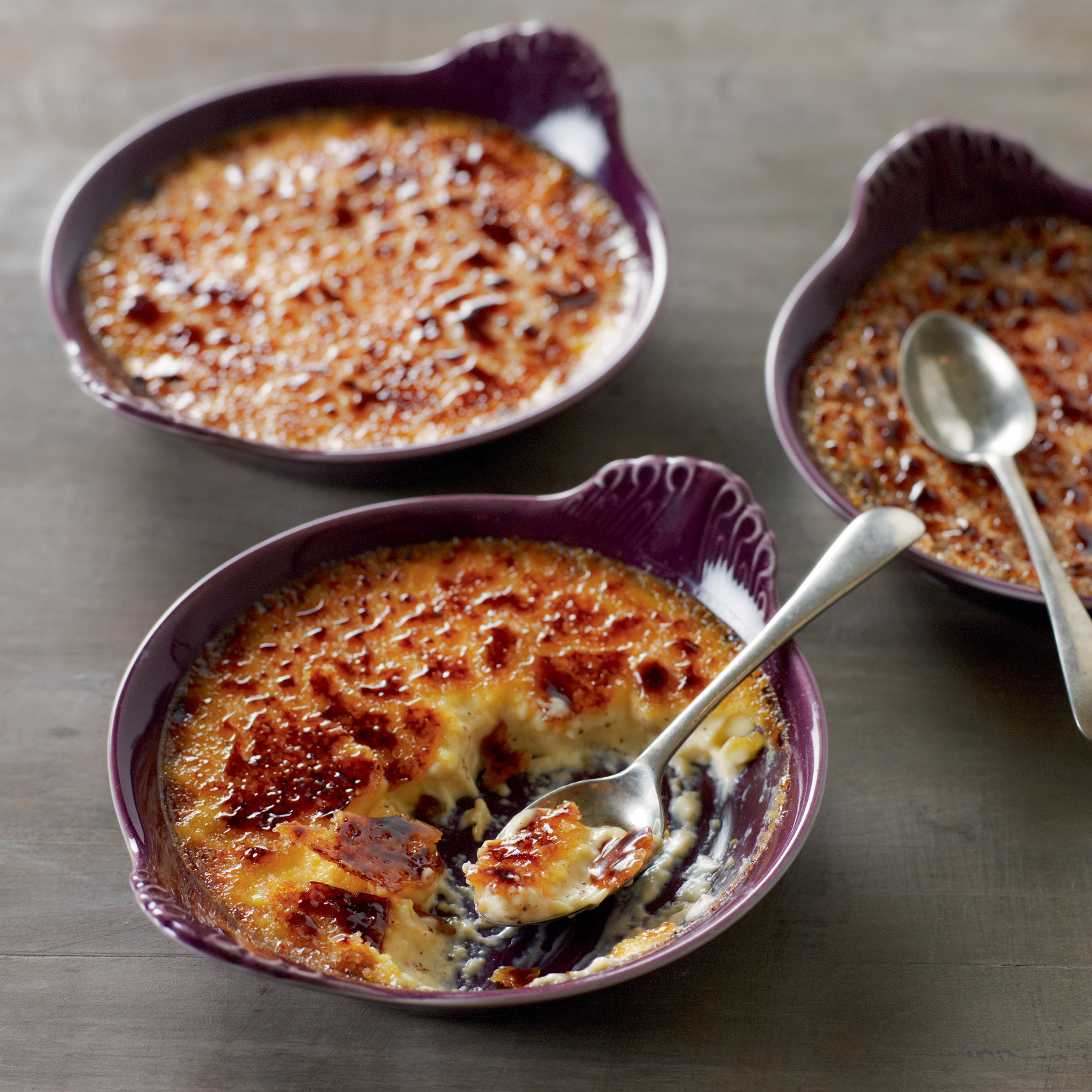 Tres ramequines morados con crema catalana, las cucharas están encima. El fondo es de madera.