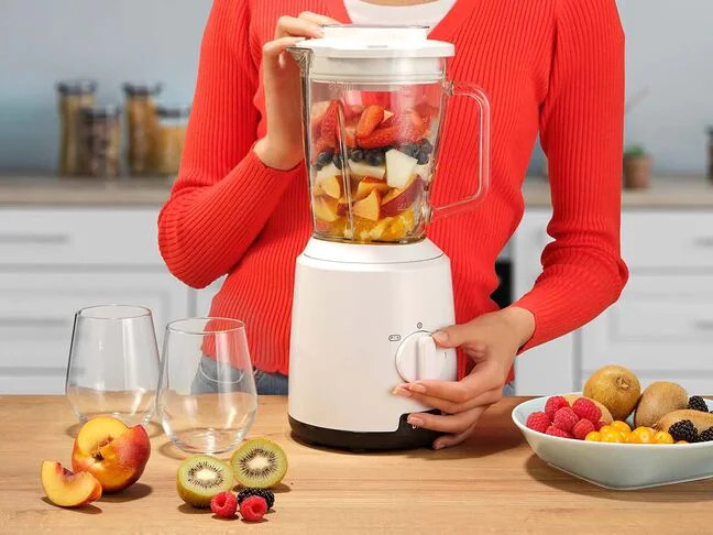 Woman using a blender. It's filled with fruit. Other fruits are beside the blender.