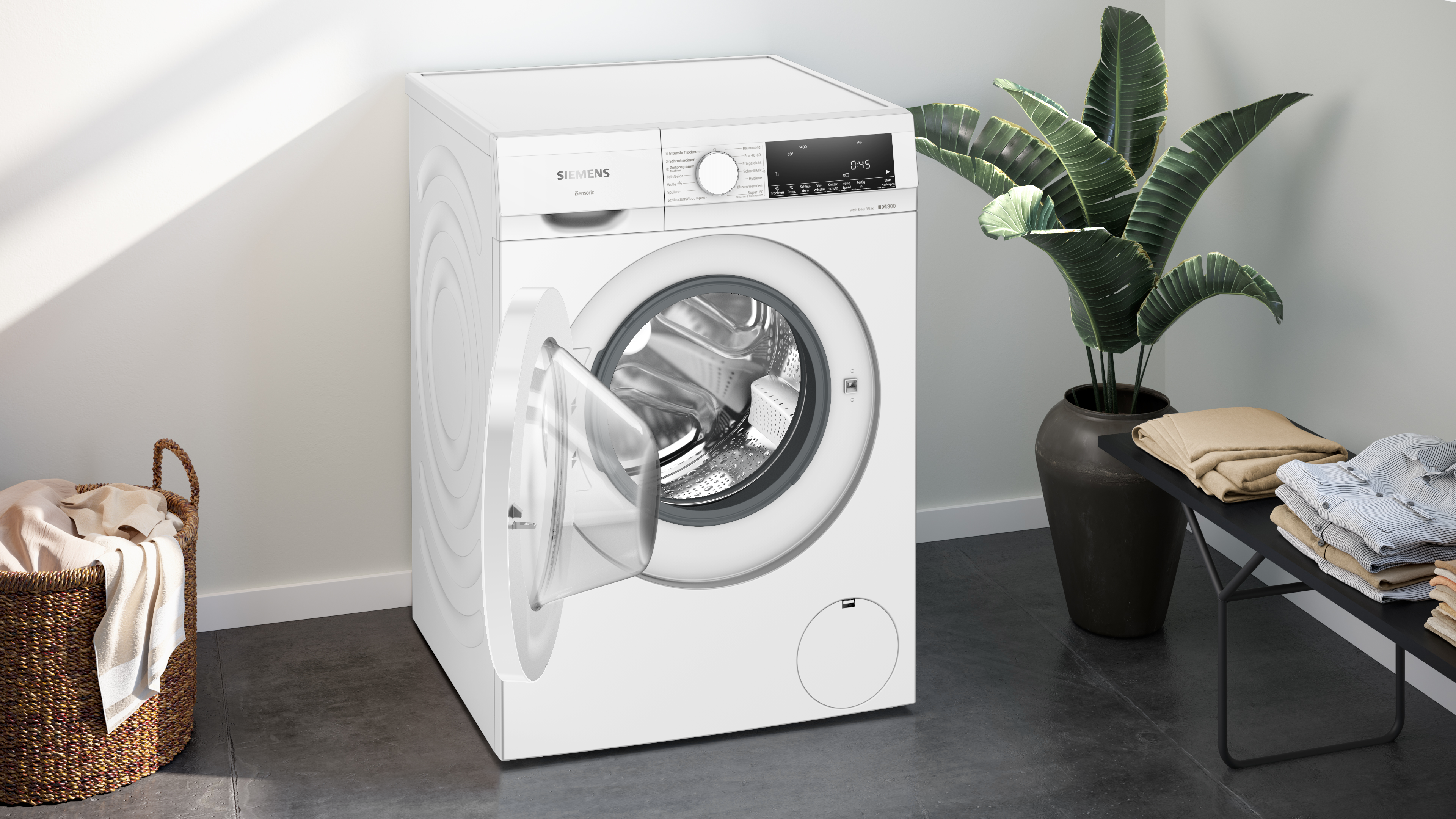 Weiße Waschmaschine mit offener Tür. Ein Korb mit Kleidung und eine Pflanze rechts.
